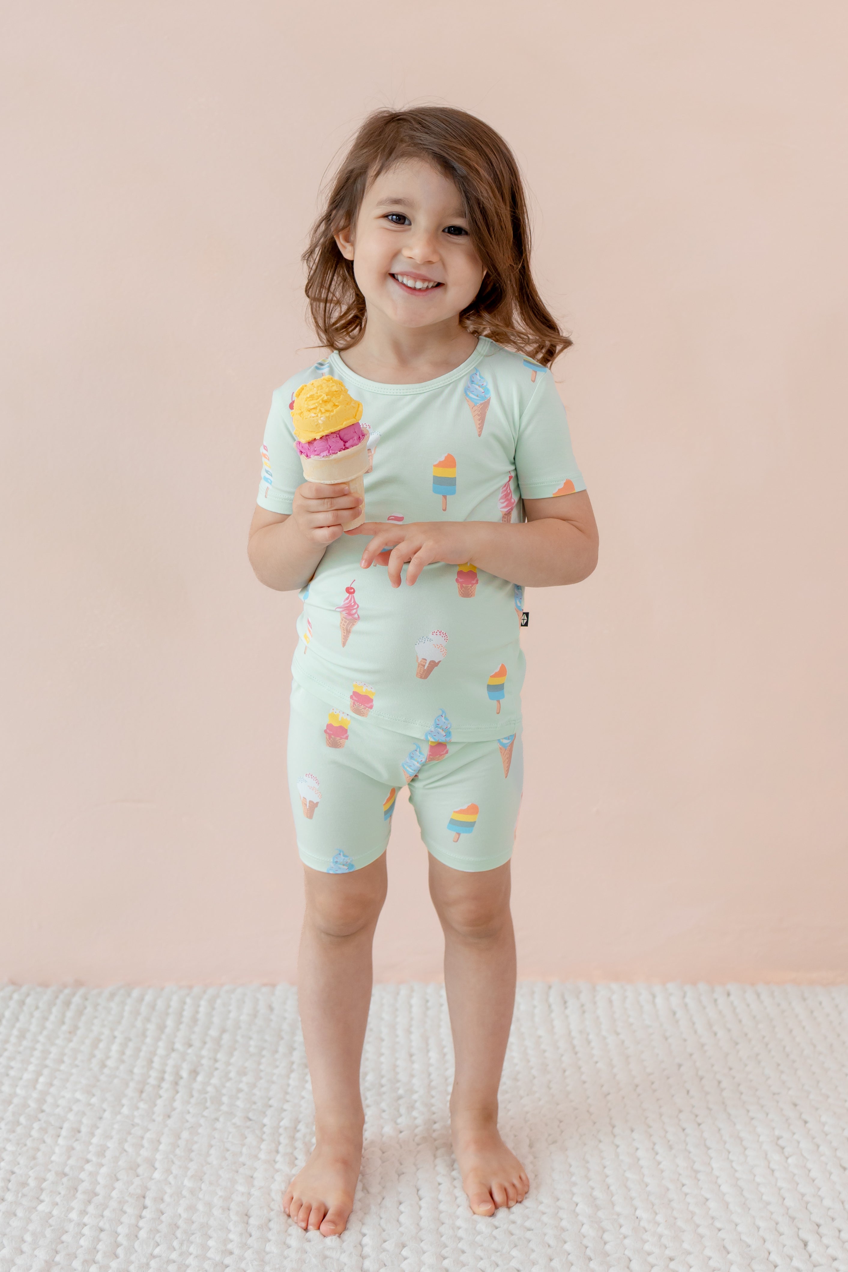 Child smiling holding an ice cream cone wearing short sleeve pajama set in Mint Ice Cream print on beige background