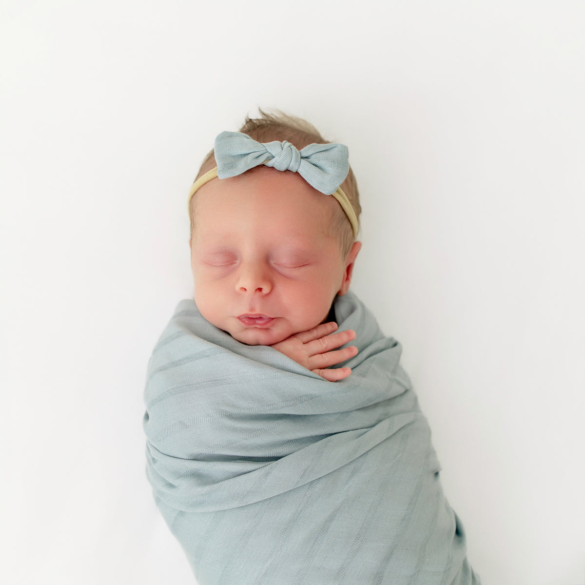 Newborn wrapped in swaddle with matching Bamboo Muslin Tiny Bow in Mist Blue
