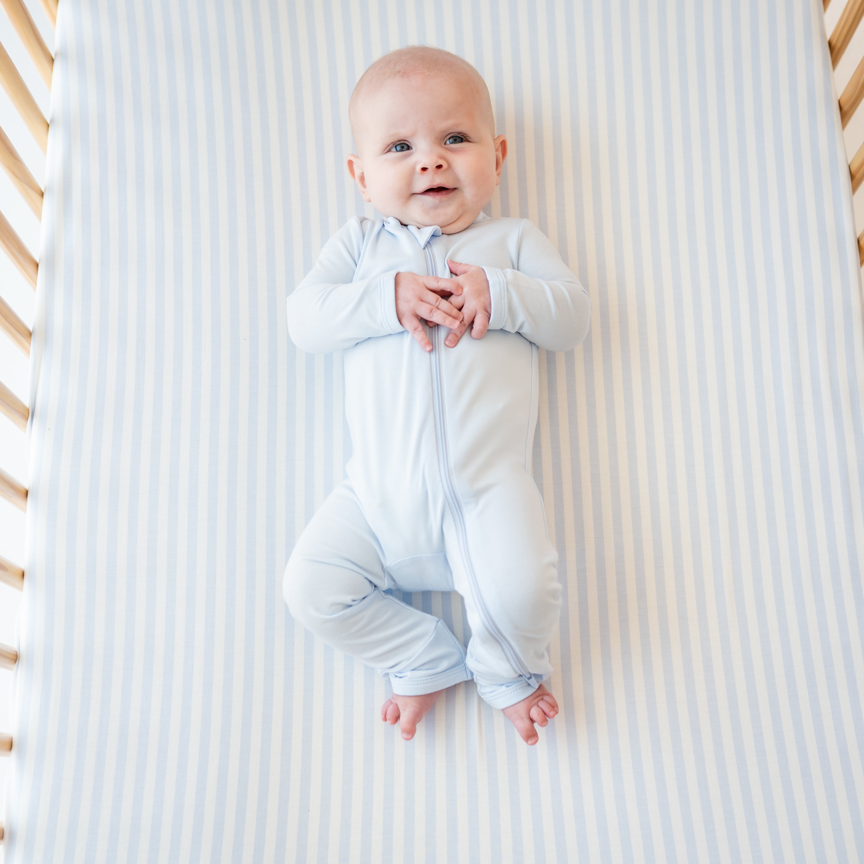 Smiling infant laying in a crib on top of a Crib Sheet in Small Mist Stripe wearing a Mist Zippered Romper