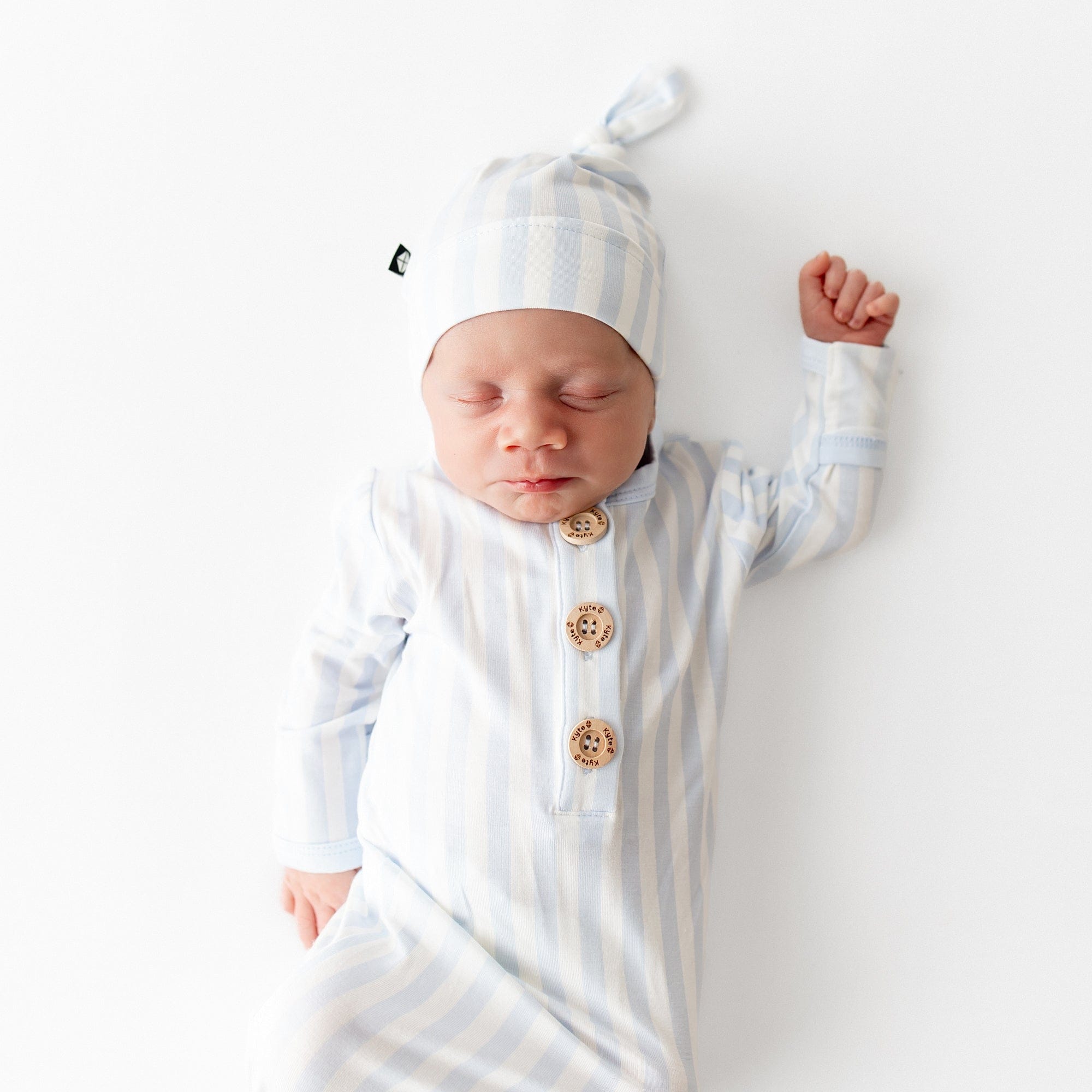 Sleeping newborn wearing the Knotted Gown with Hat Set in Small Mist Stripe laying on a white surface