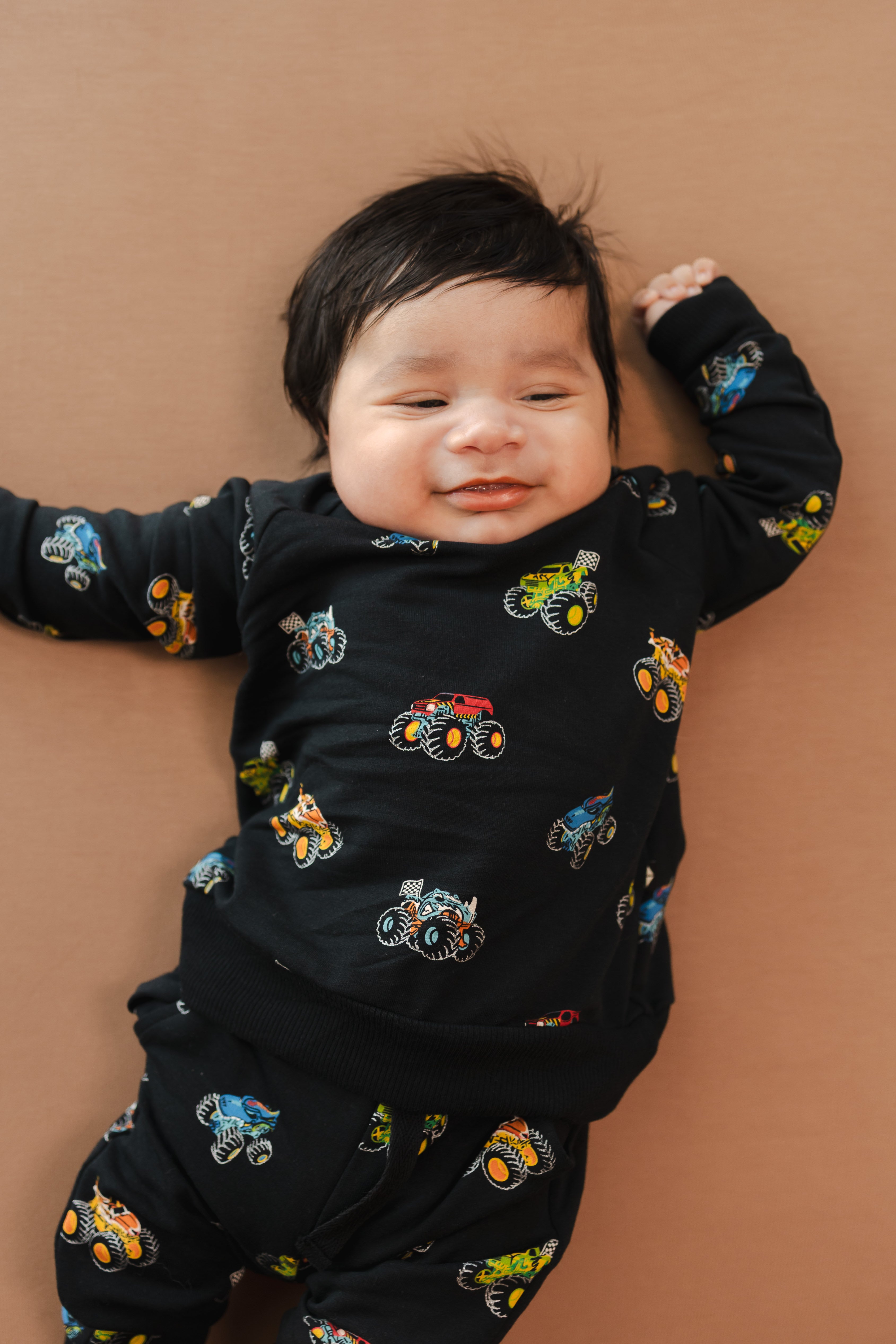 Baby smiling in black Long Sleeve Jogger Set in Monster Truck print on beige background