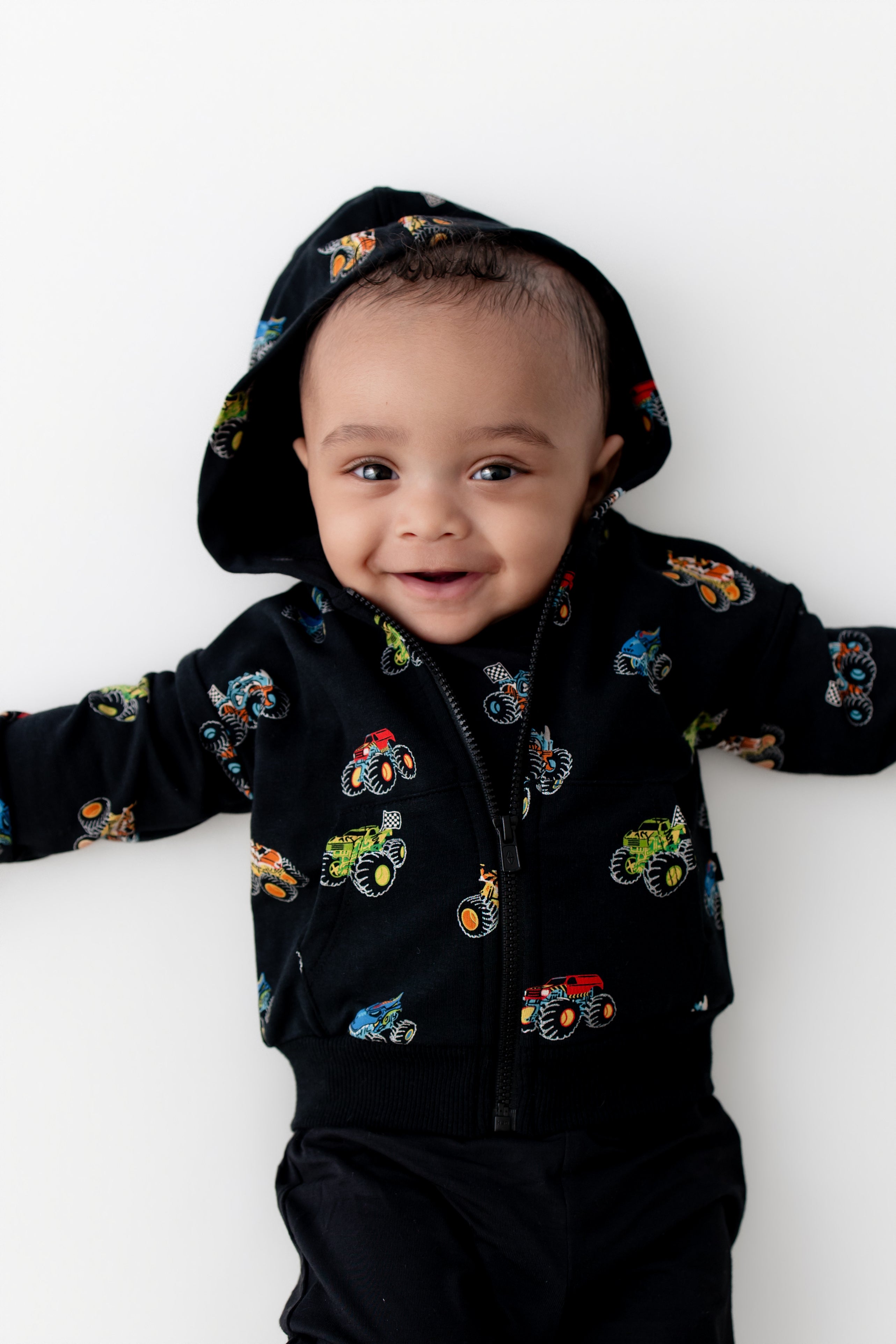 Baby in Black Zip-Up Hoodie in Monster Truck print on black pants on a white background