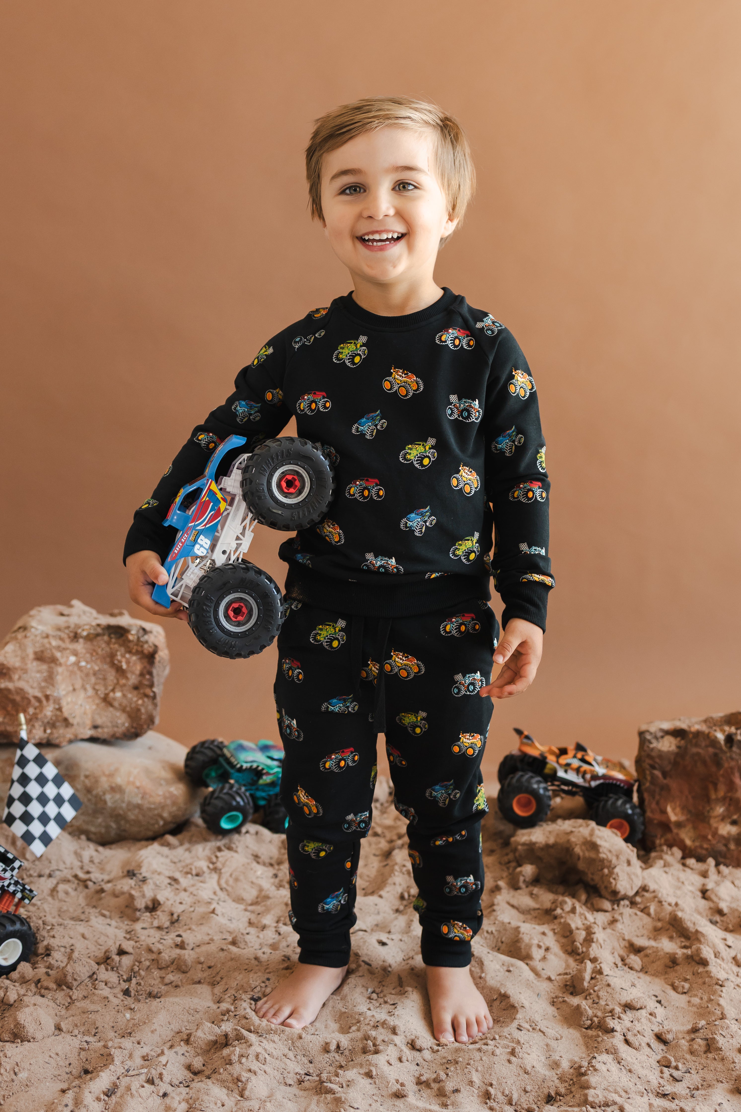 Toddler wearing Black Long Sleeve Jogger Set in Monster Truck print holding a truck standing on sand and rock setup