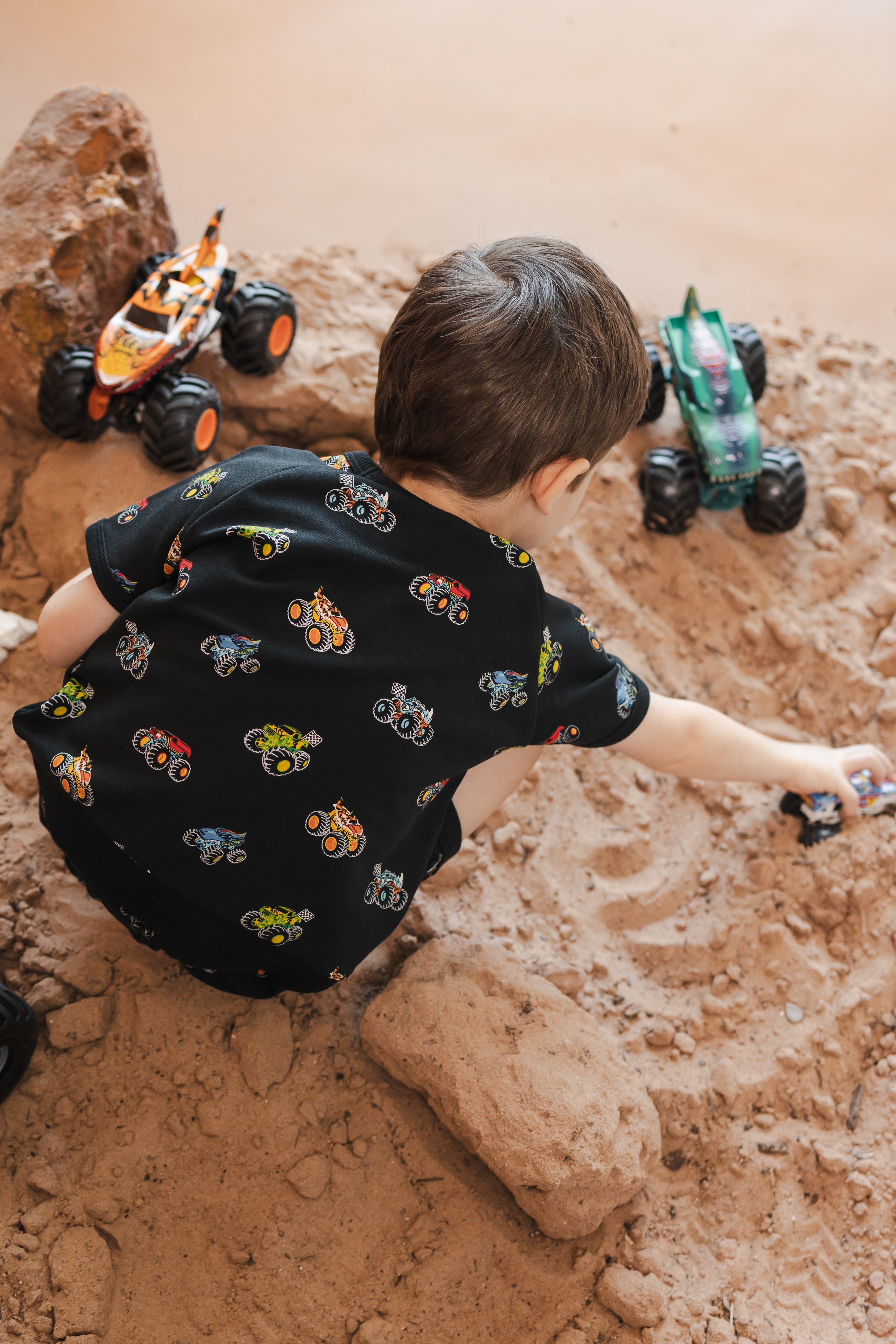 Boy playing with toy truck wearing Black Short Sleeve Jogger Set in Monster Truck print on sand and rocks with toy trucks.