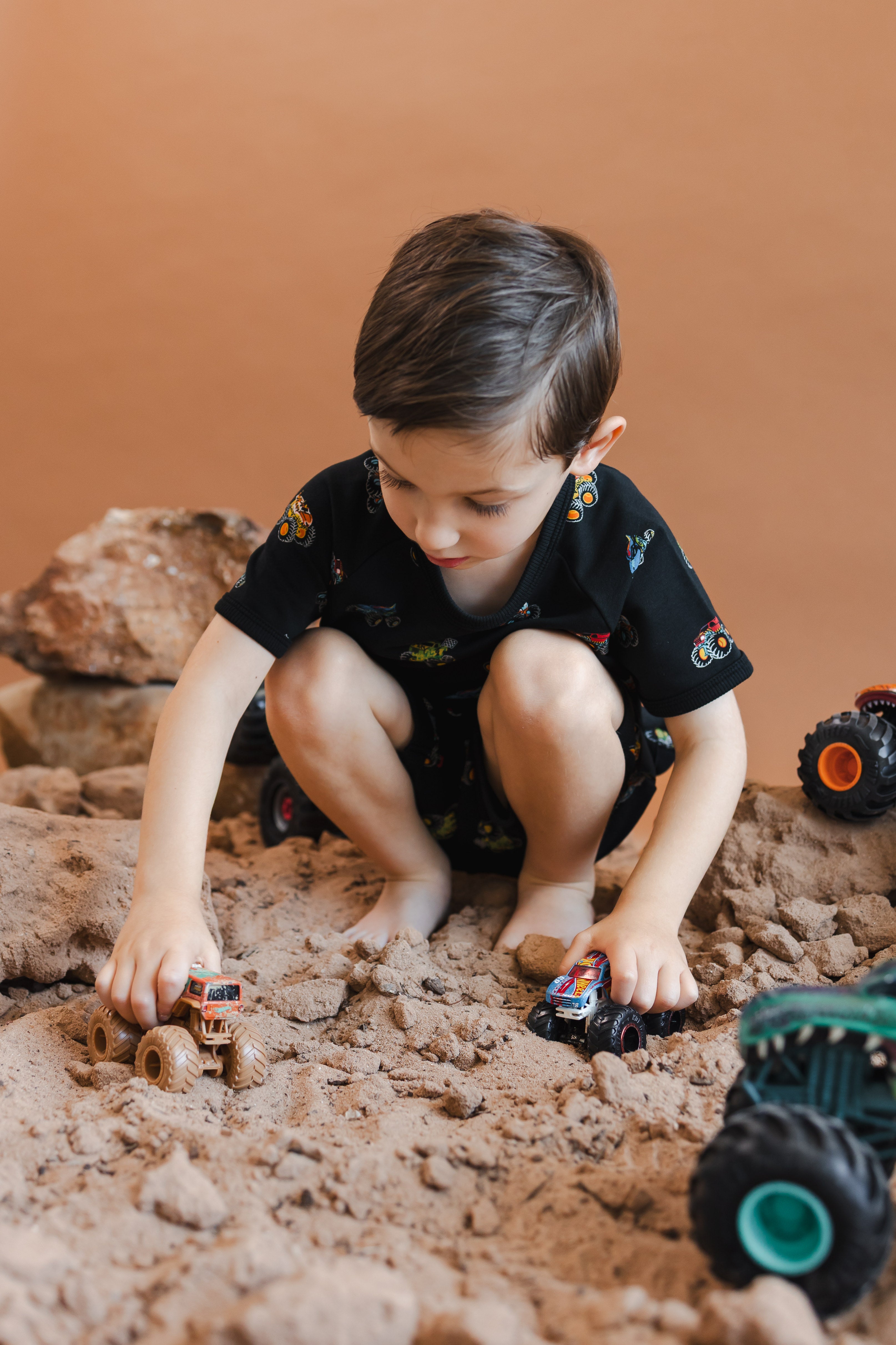 Boy playing with toy truck on sand and rocks while wearing Black Short Sleeve Jogger Set in Monster Truck print