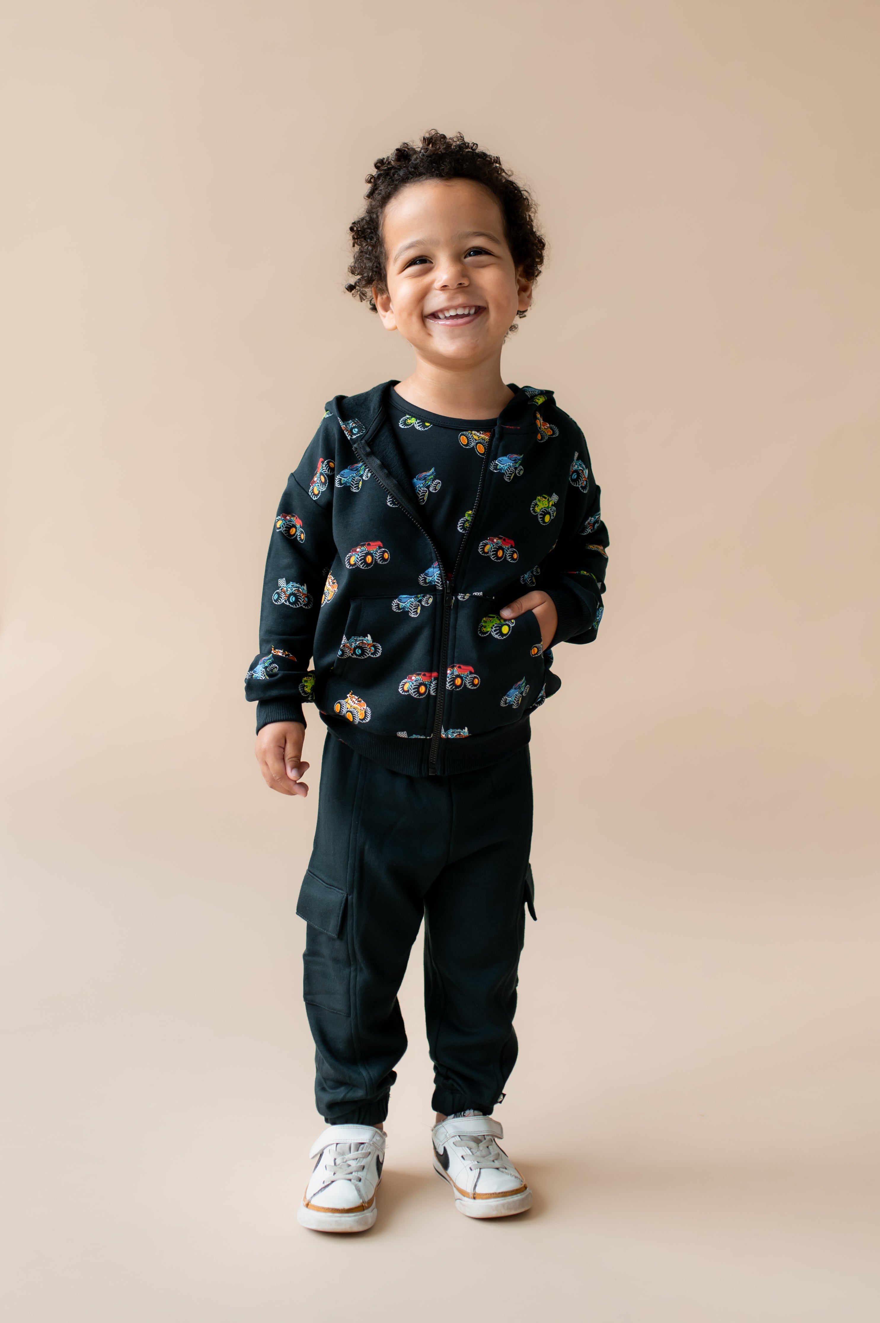Boy smiling in Black Zip-Up Hoodie in Monster Truck print and black pant and white shoes on a beige background
