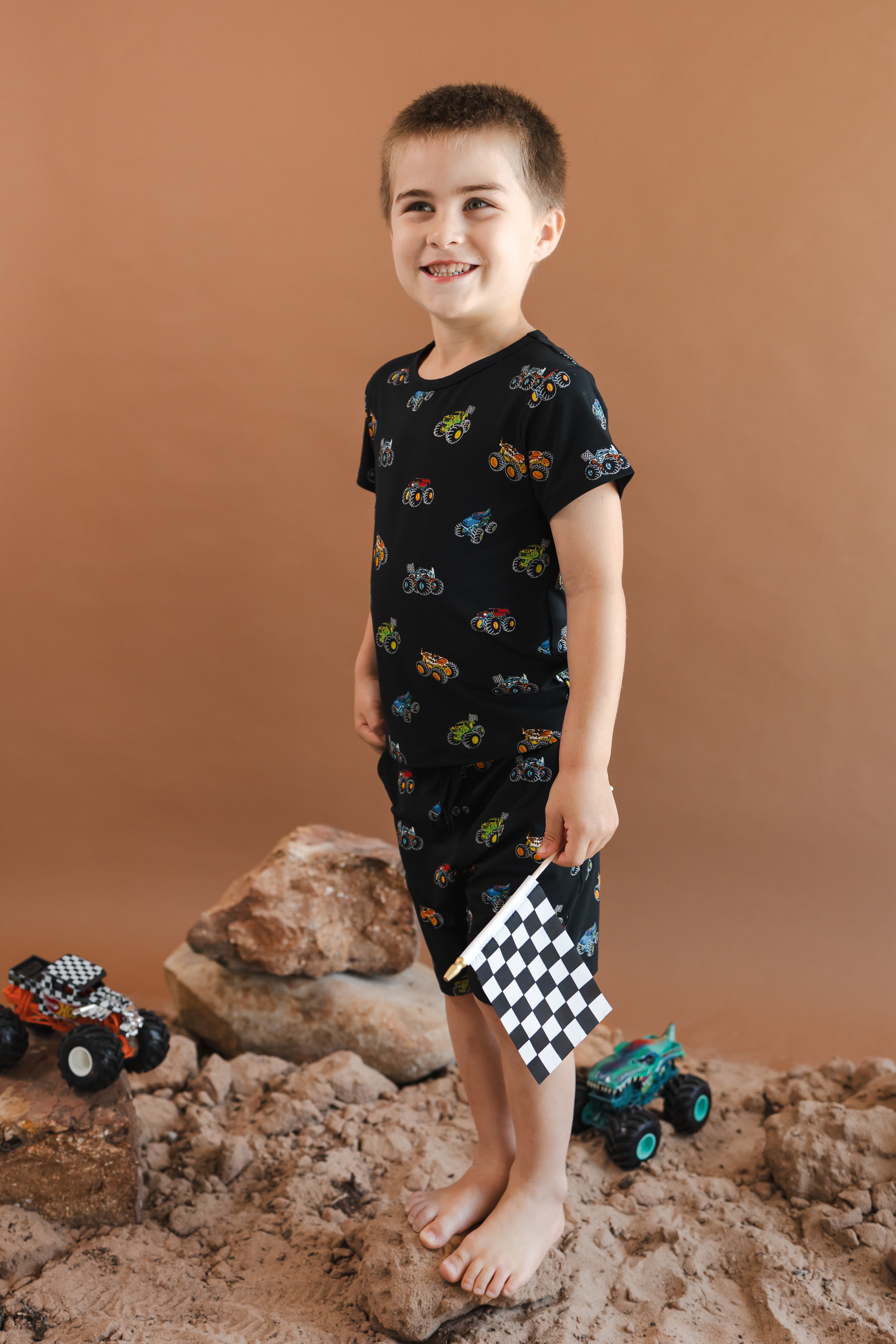 Boy smiling in Black Toddler Basic Tee and drawstring short in Monster Truck print standing on sand and rock with toy trucks