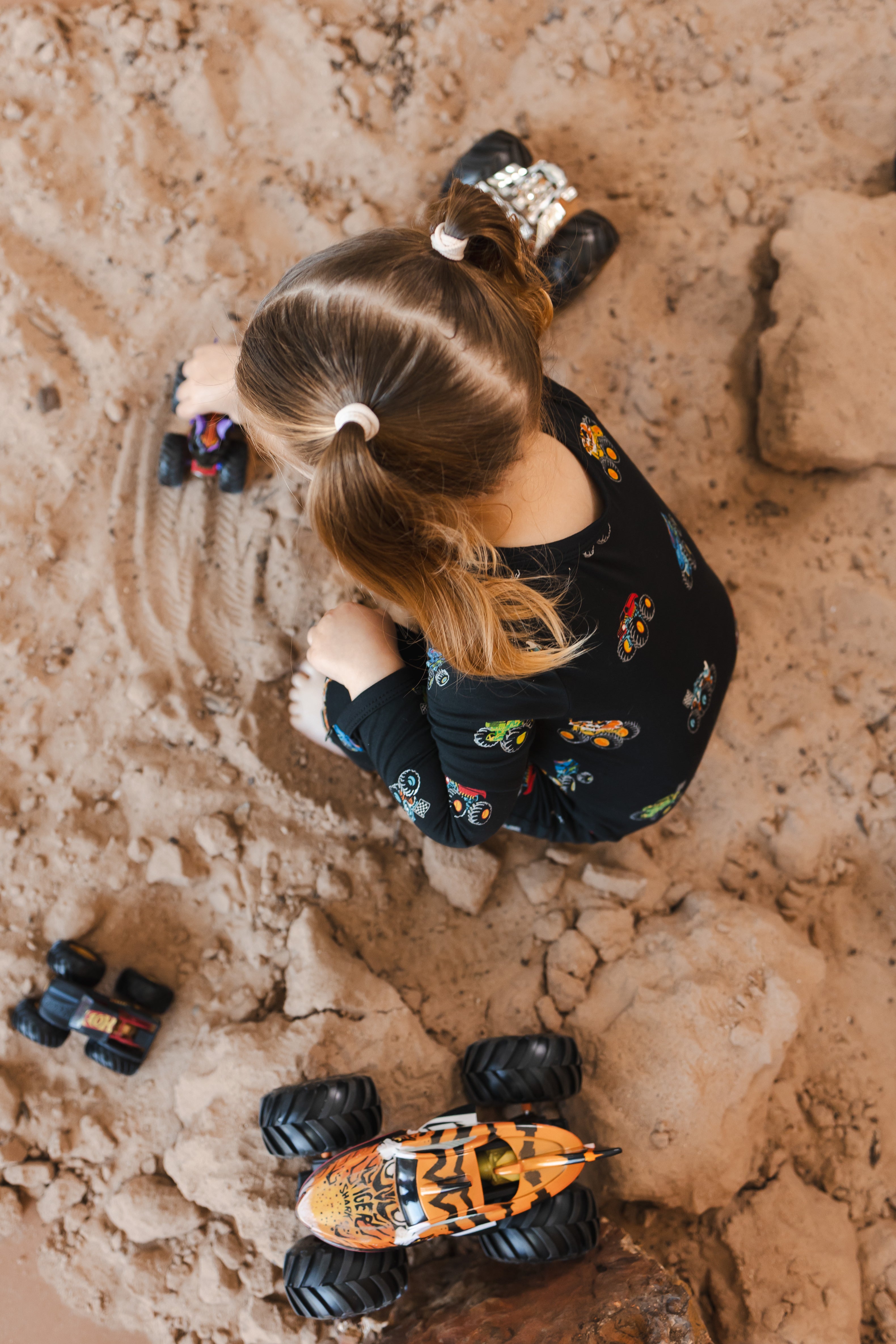 Girl wearing black Zippered Romper in Monster Truck print playing with toy trucks on sand and rocks