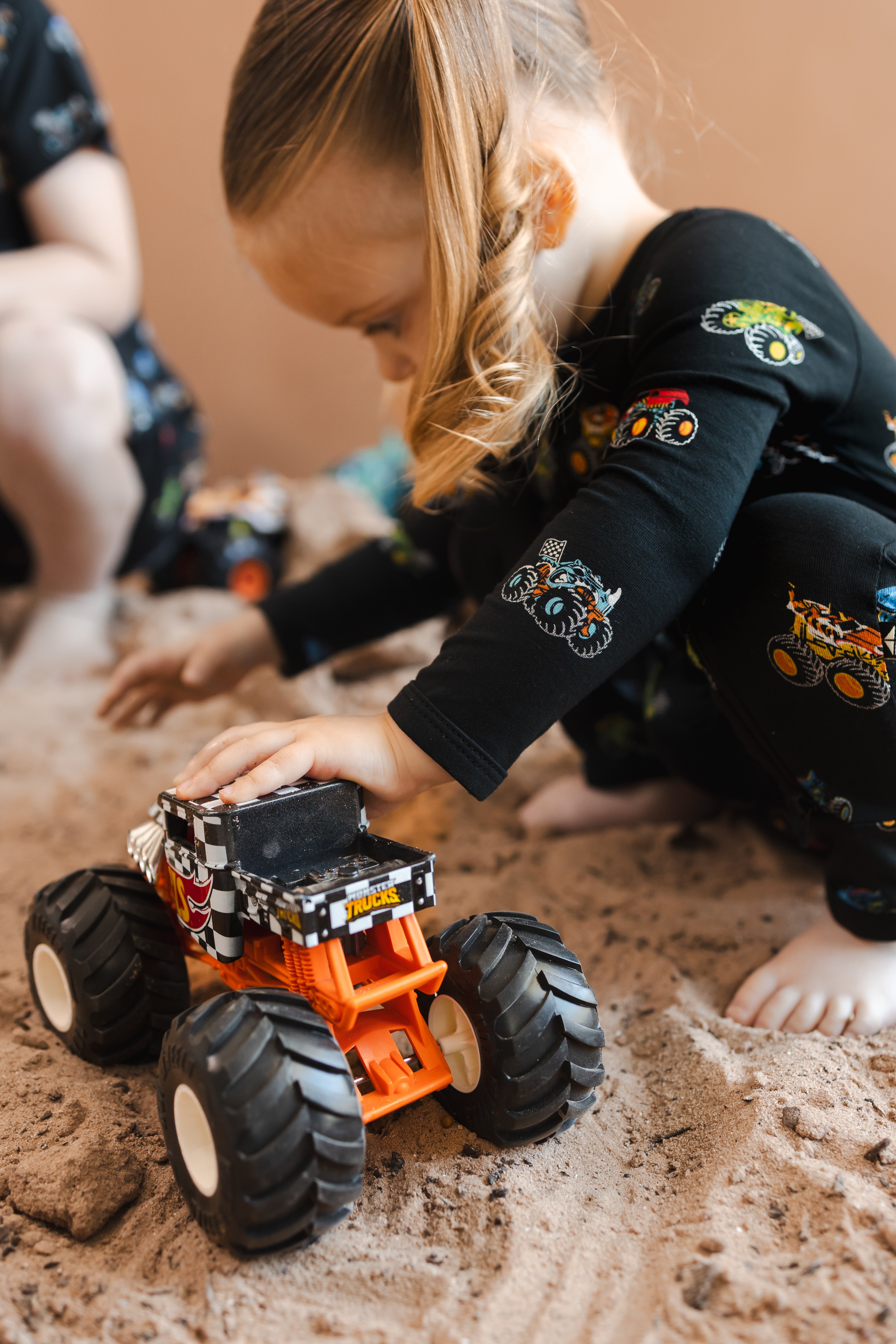 Girl wearing black Zippered Romper in Monster Truck print sitting on sand and rocks playing with toy truck
