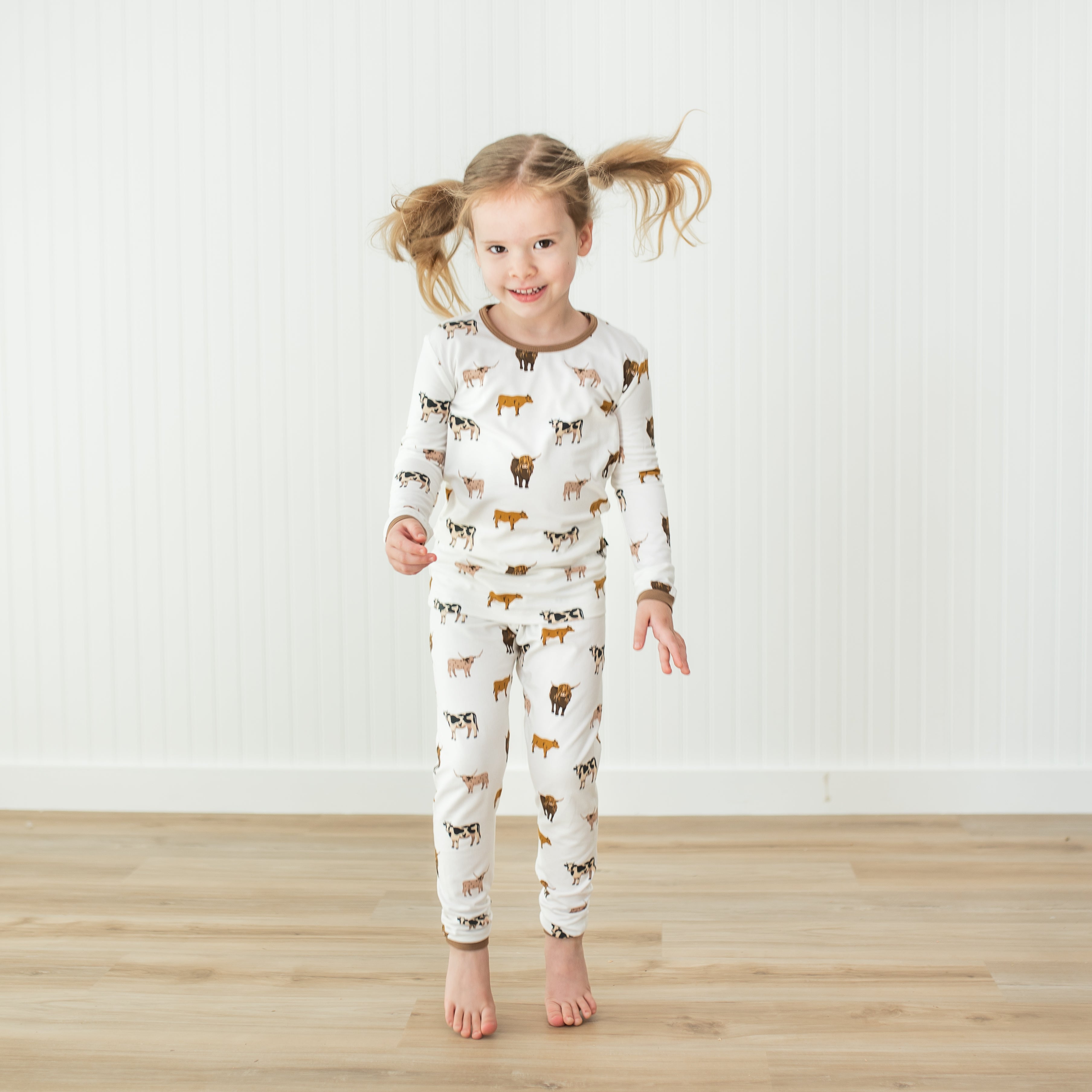 Young girl wearing a white pajama set with cow prints on a wooden floor.