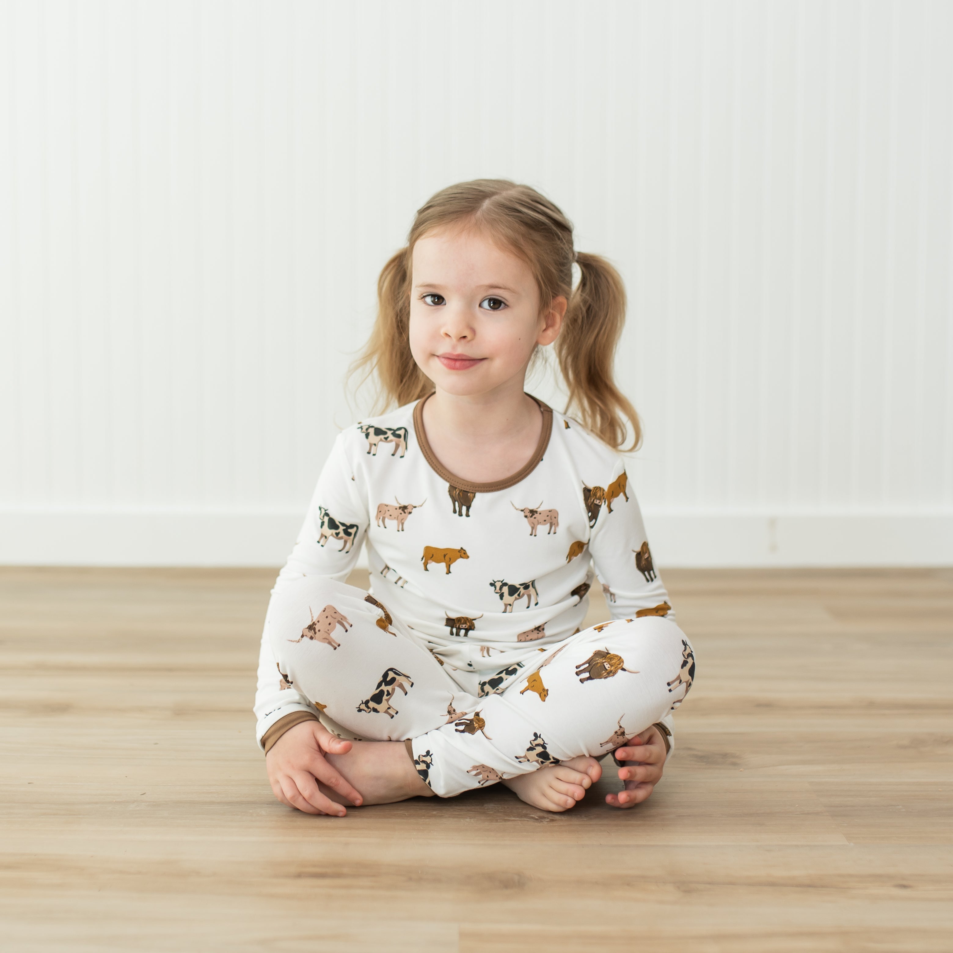 Child wearing pajamas with cow prints sitting on a wooden floor.