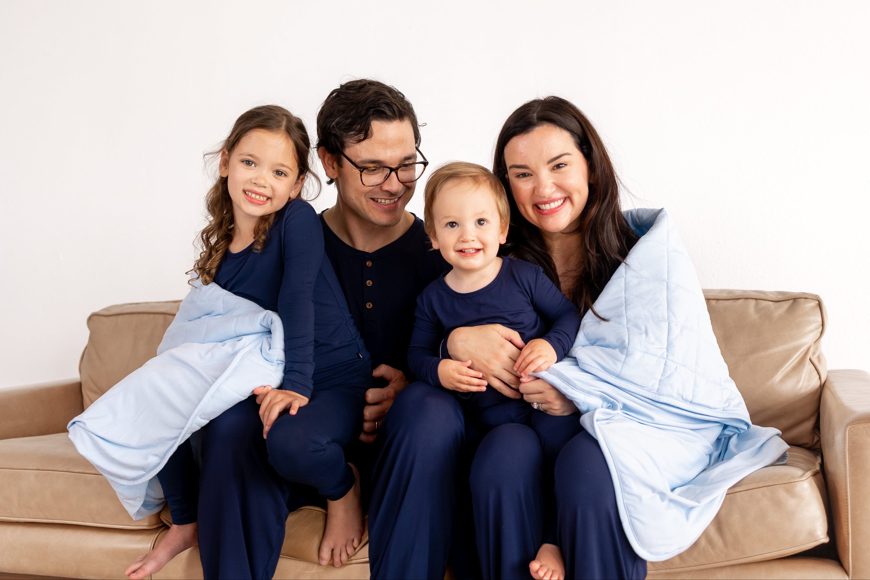 Family of 4 in matching navy pajamas wrapped in a light blue bamboo blanket sitting on couch