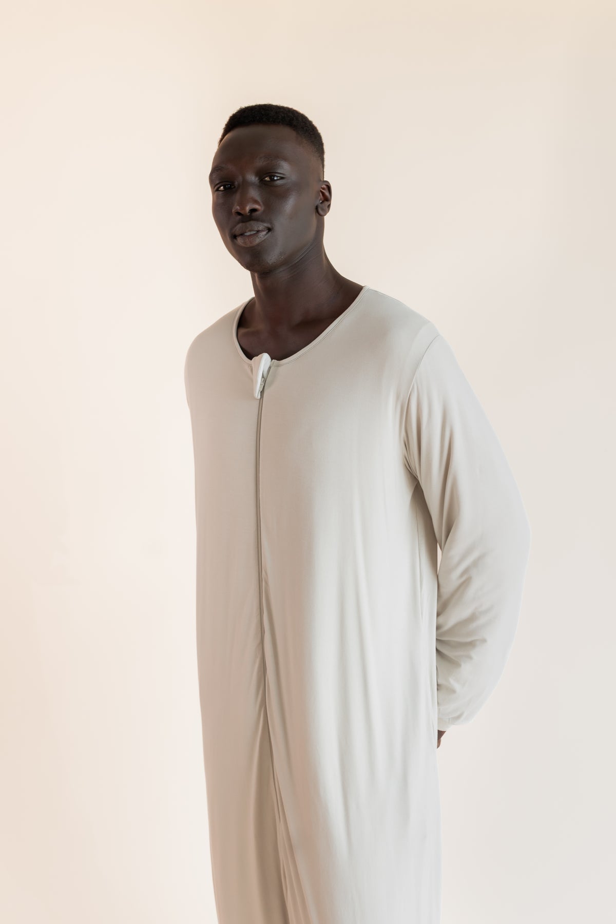 A man wearing a long-sleeved, full-length cream-colored sleep sack , standing against a solid light beige background.