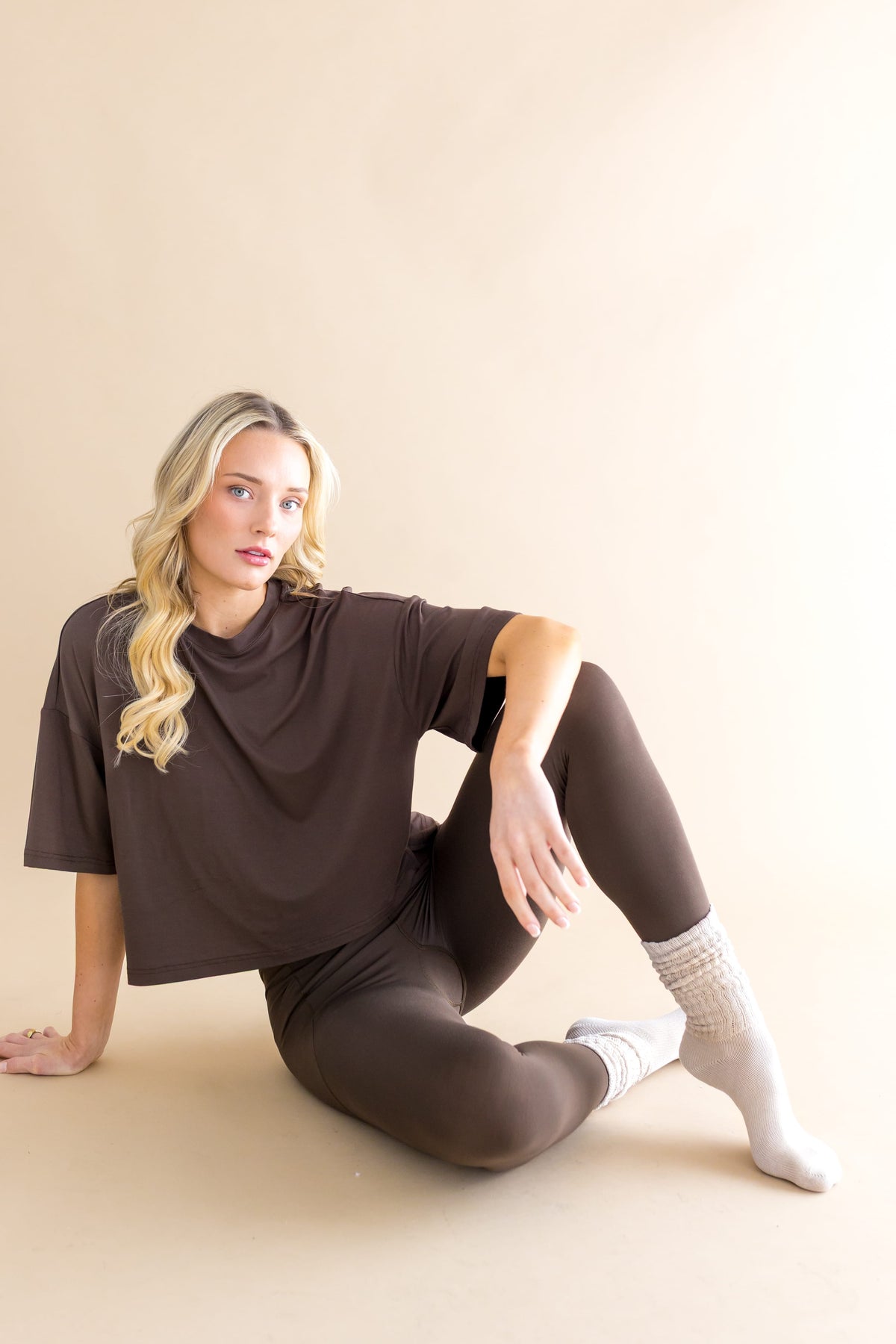 Female model sitting on the floor wearing the Adult Scrunch Socks in Oat with dark brown leggings and shirt