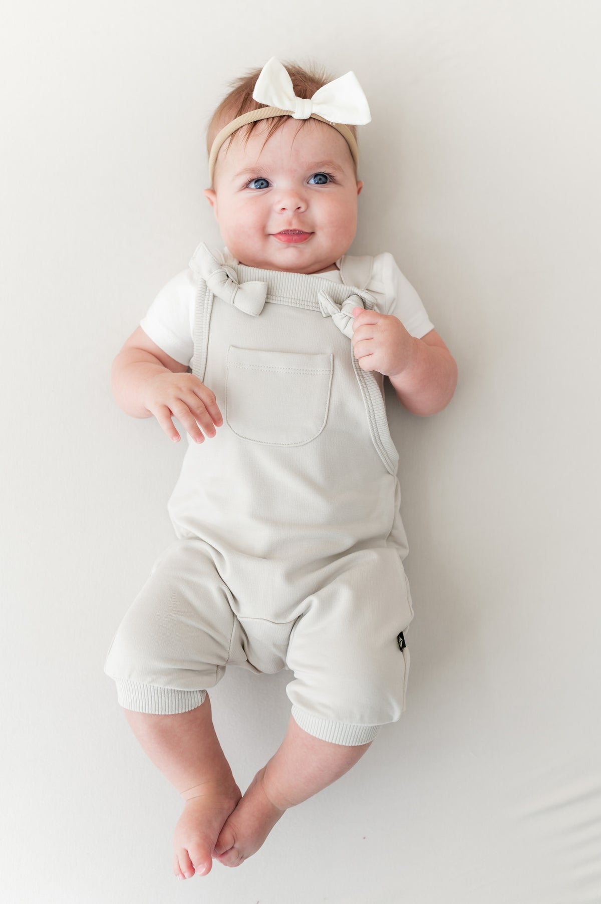 Baby wearing Overall in Oat color with white bow headband on a beige background
