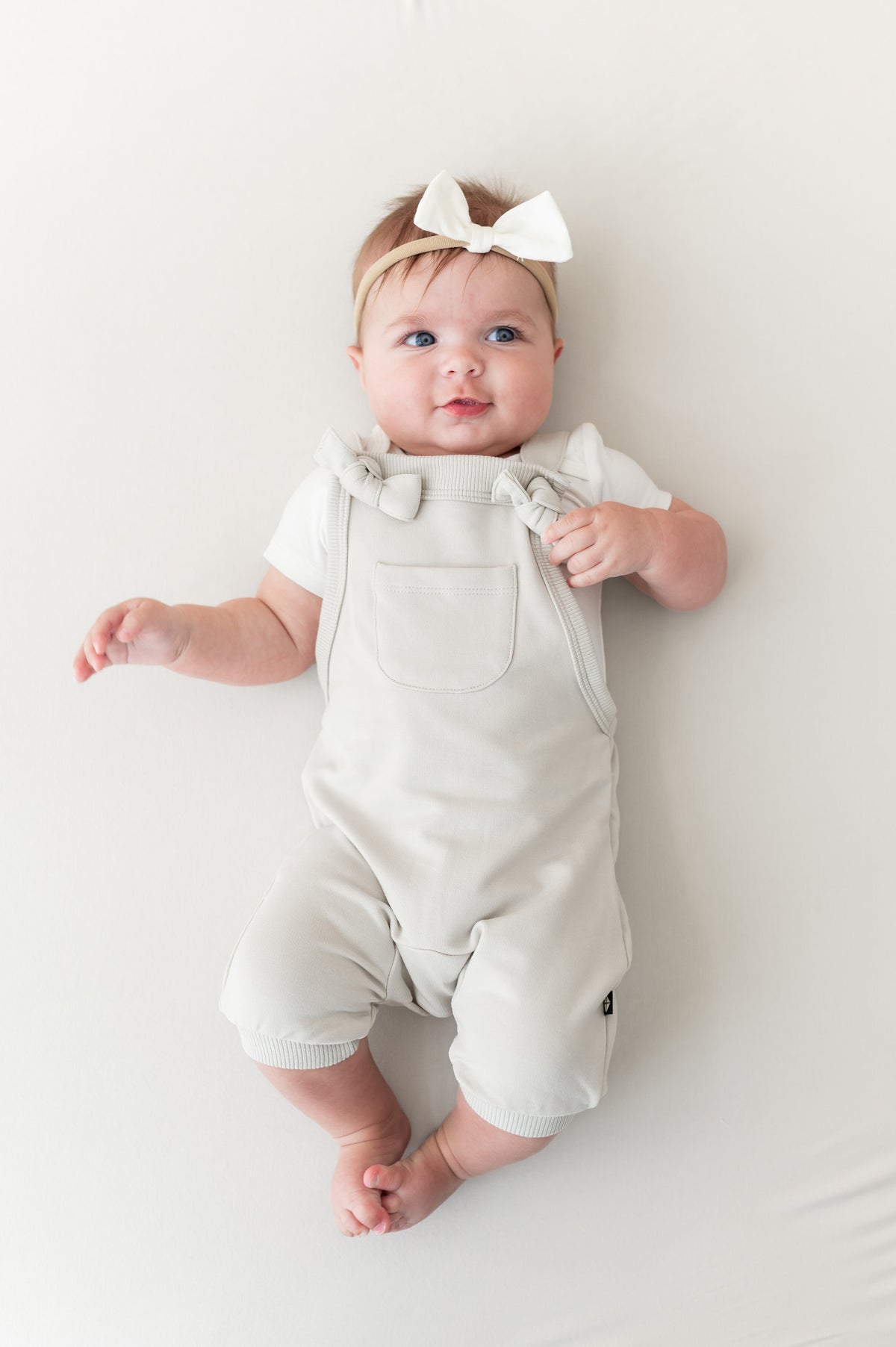 Baby wearing Overall in Oat color with white bow headband lying on a beige background