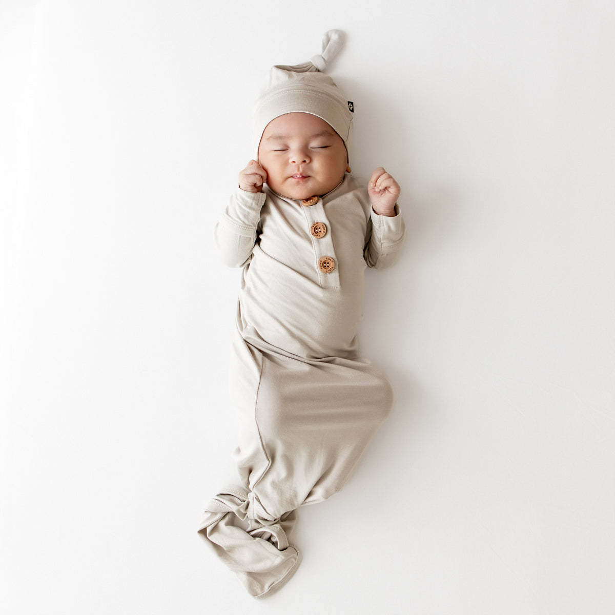 Baby in oat colored knotted gown outfit that has wooden buttons and a matching hat