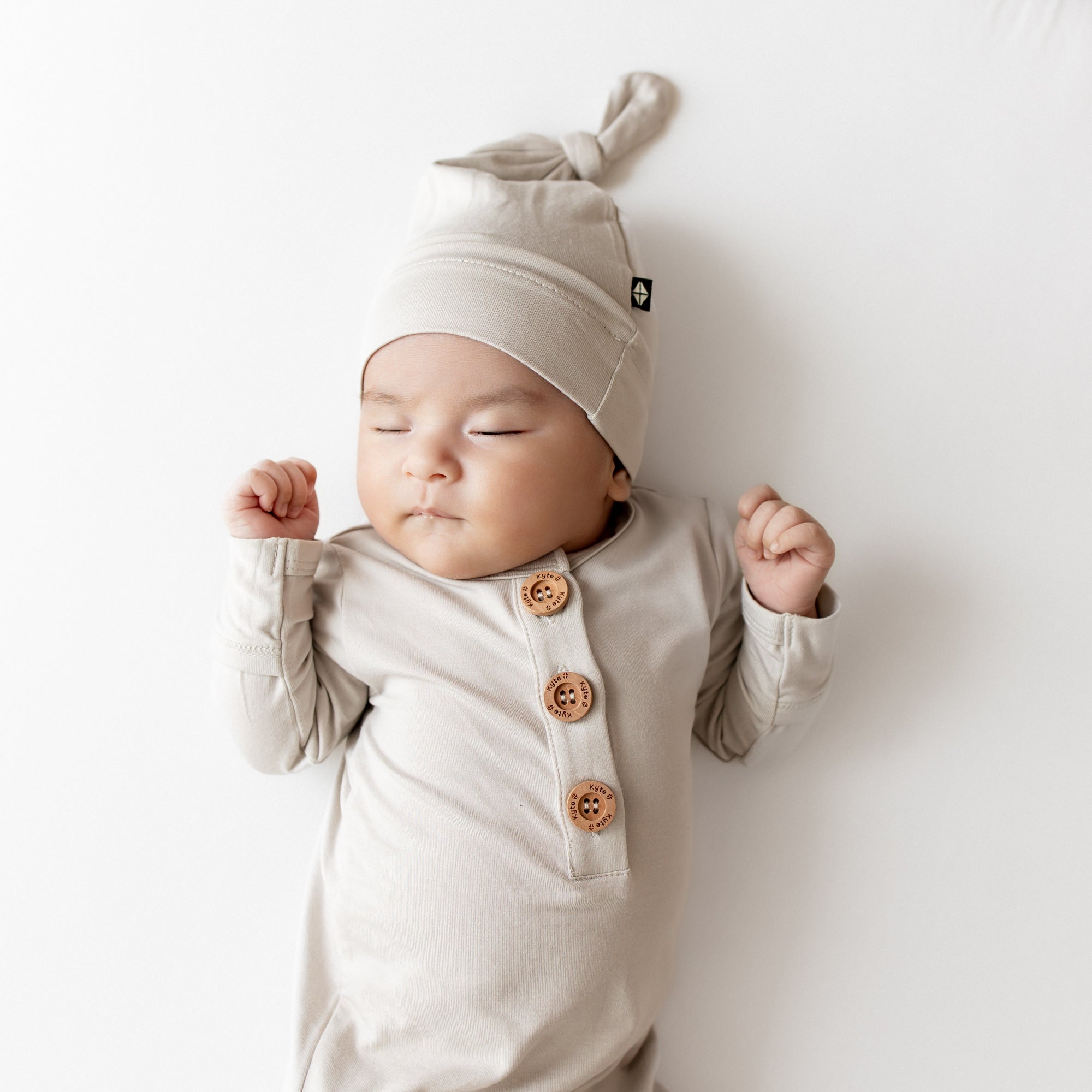 Close up of Baby in oat colored knotted gown outfit that has wooden buttons and a matching hat