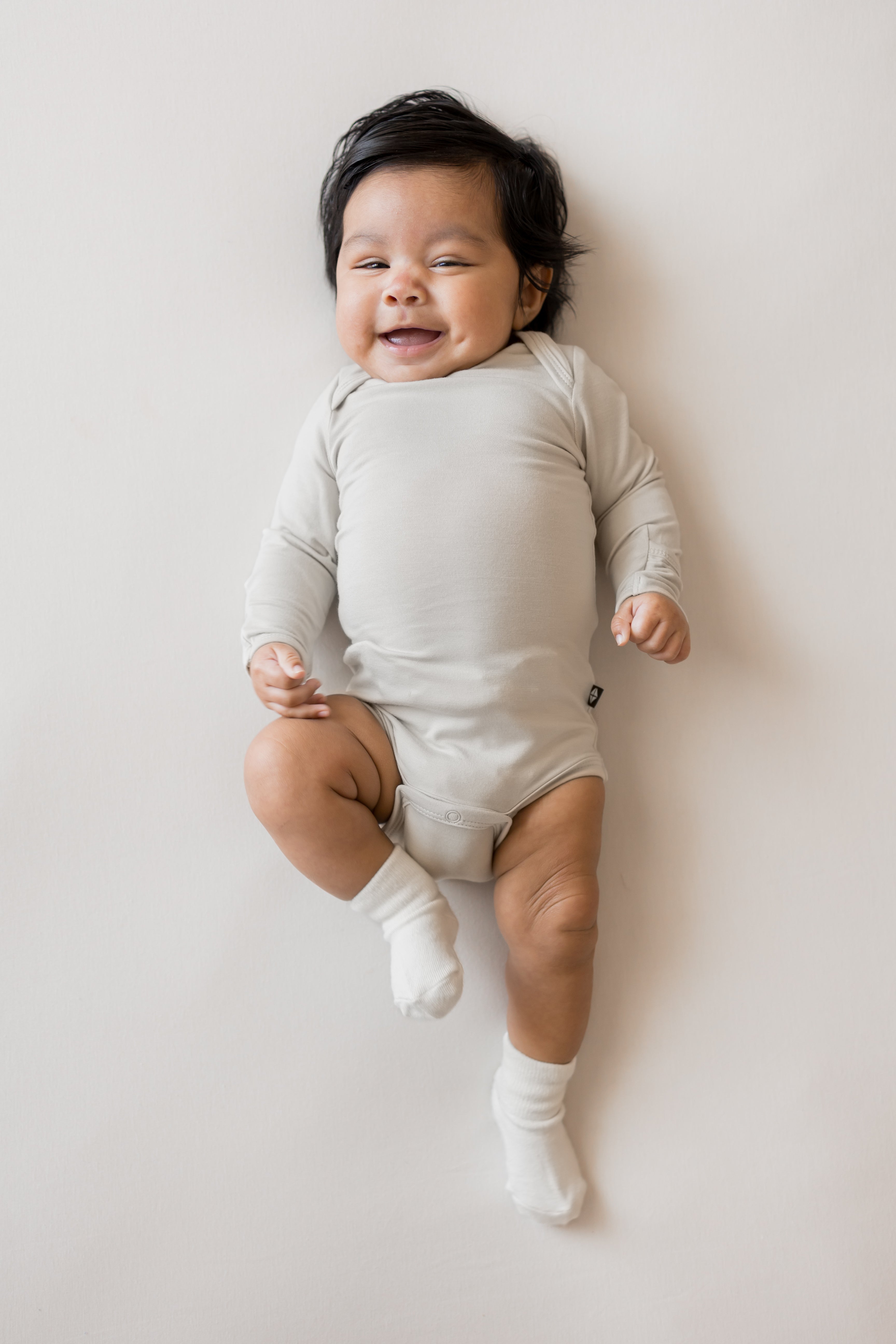 Baby smiling in Long Sleeve Bodysuit in Oat color and white socks on a beige background