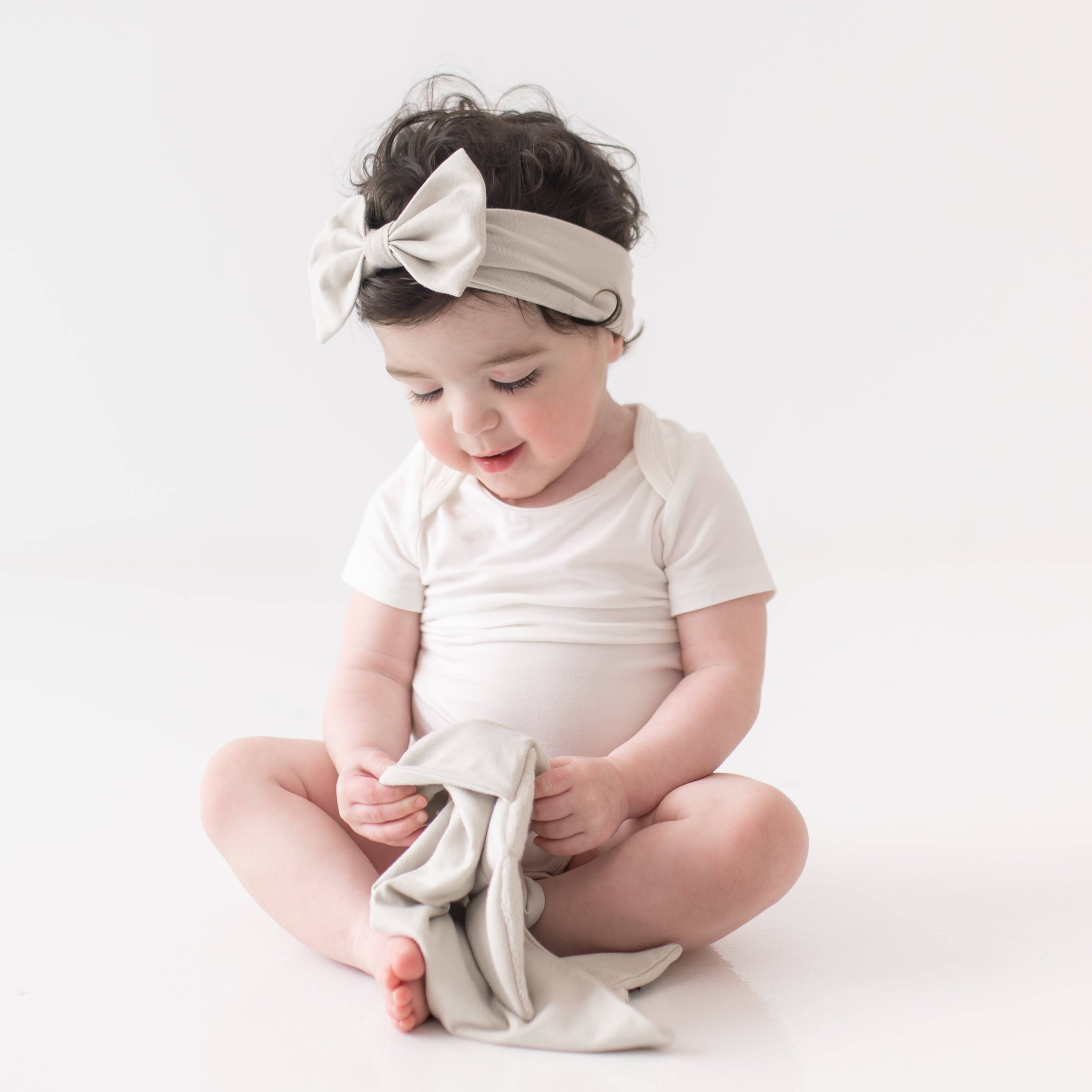 Baby wearing Bow Headband in Oat and white bodysuit while playing with Kite Lovey in oat