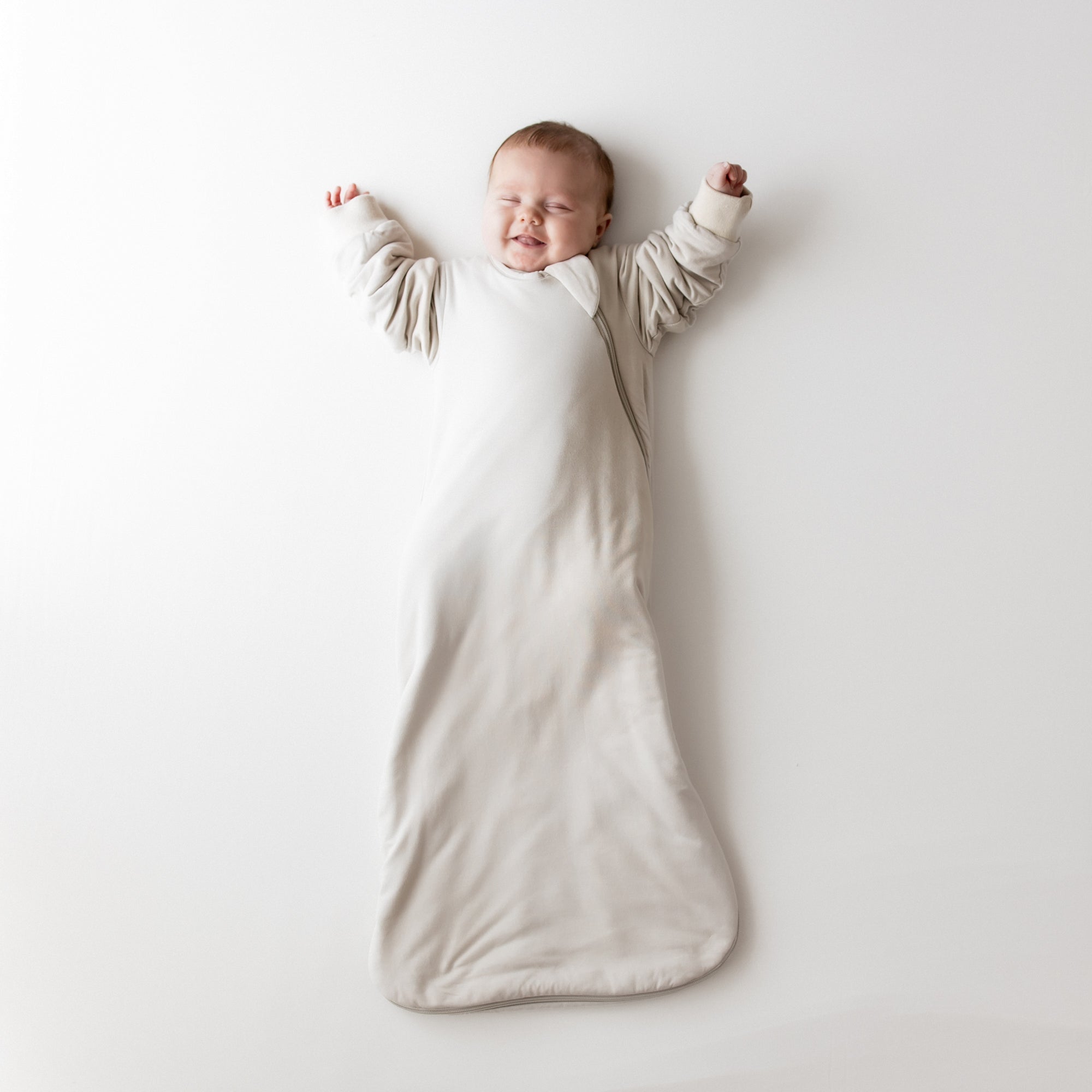 Baby in oat slumber bag with arms raised on a neutral backdrop