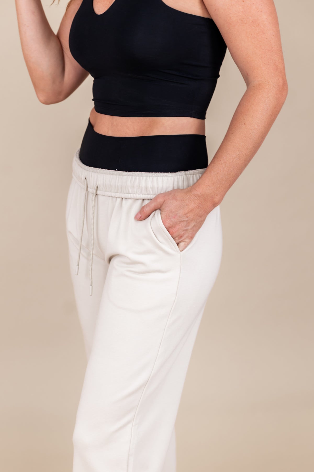 Woman wearing Women's Sweatpants in Oat color with pockets under black flex bra tank on a beige background