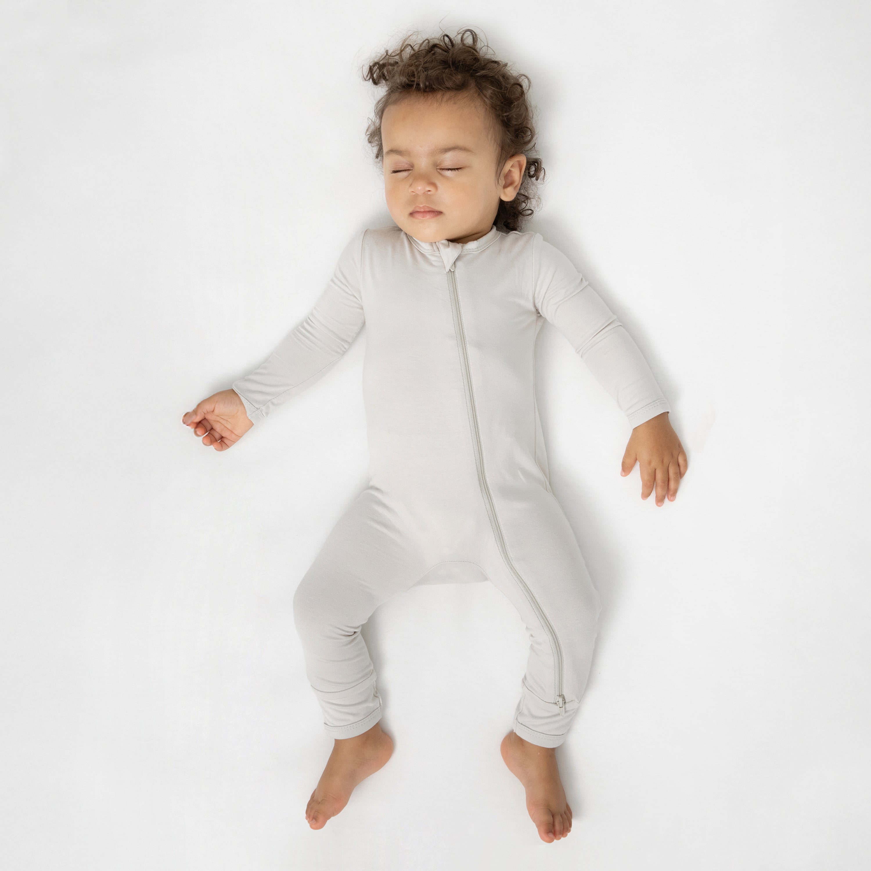 Sleeping toddler laying in a crib on a white crib sheet wearing the Zippered Romper in Oat