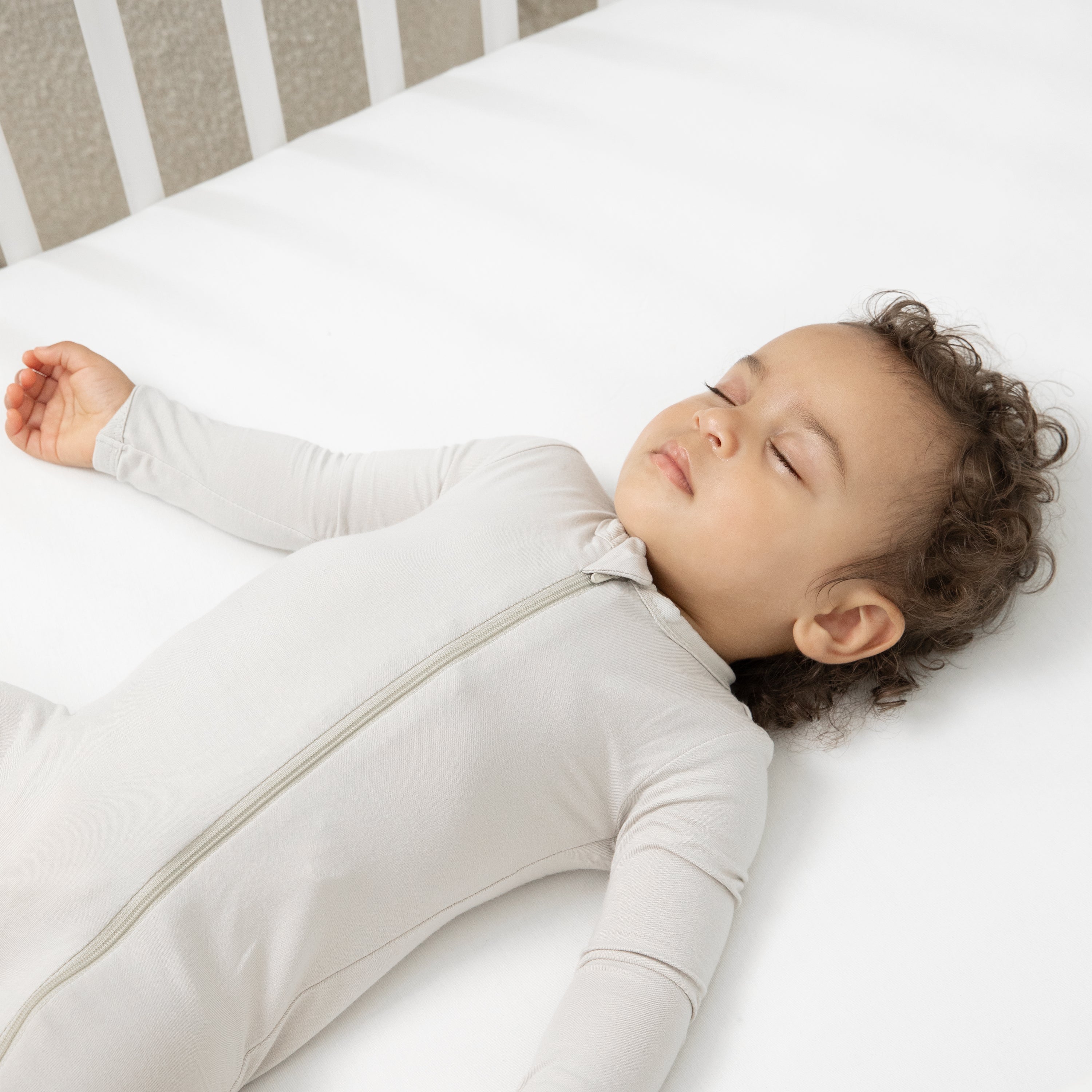 Close up of a sleeping toddler laying in a crib wearing the Zippered Romper in Oat