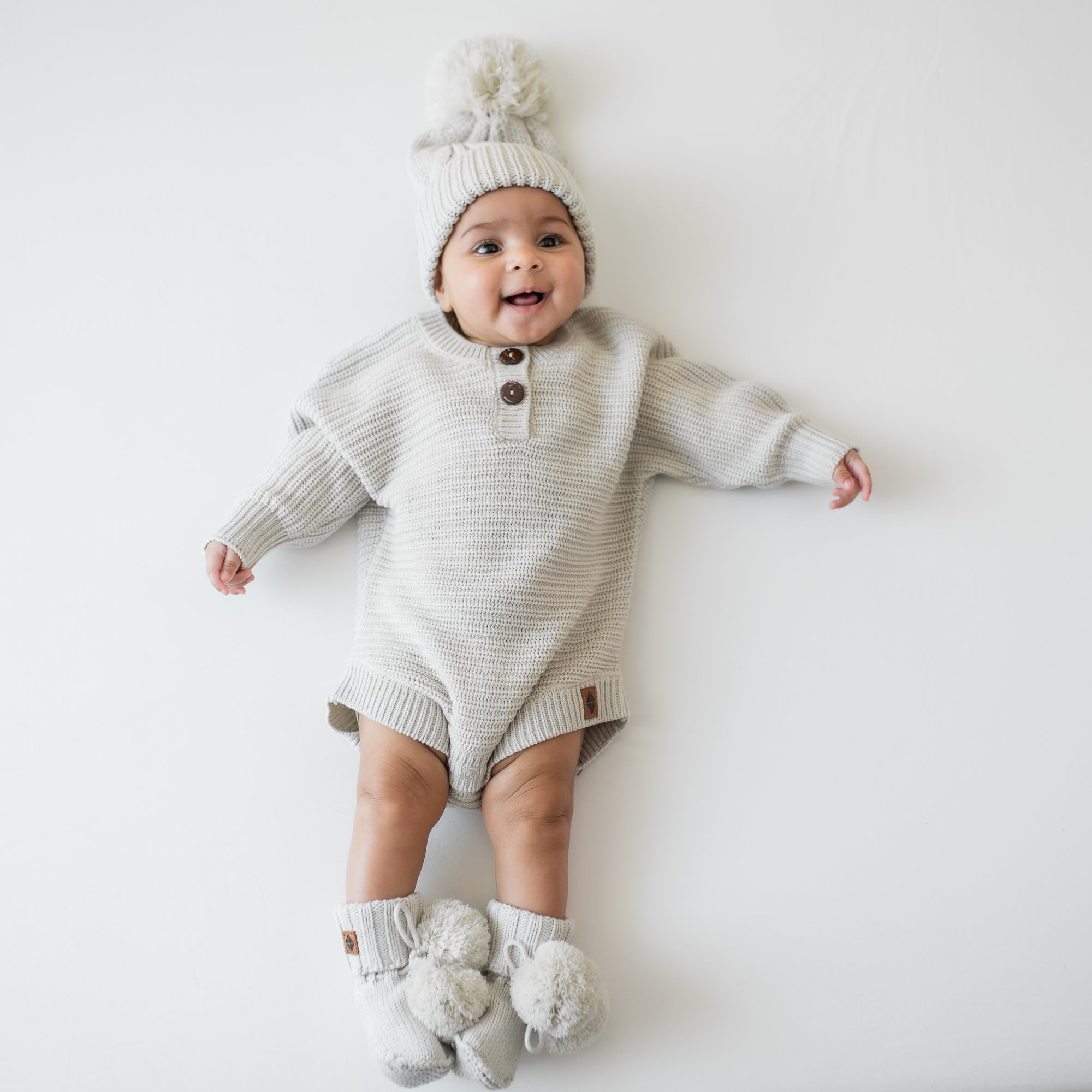 Infant laying down wearing the Chunky Cable Knit Beanie in Oat and matching chunky knit romper and knit booties