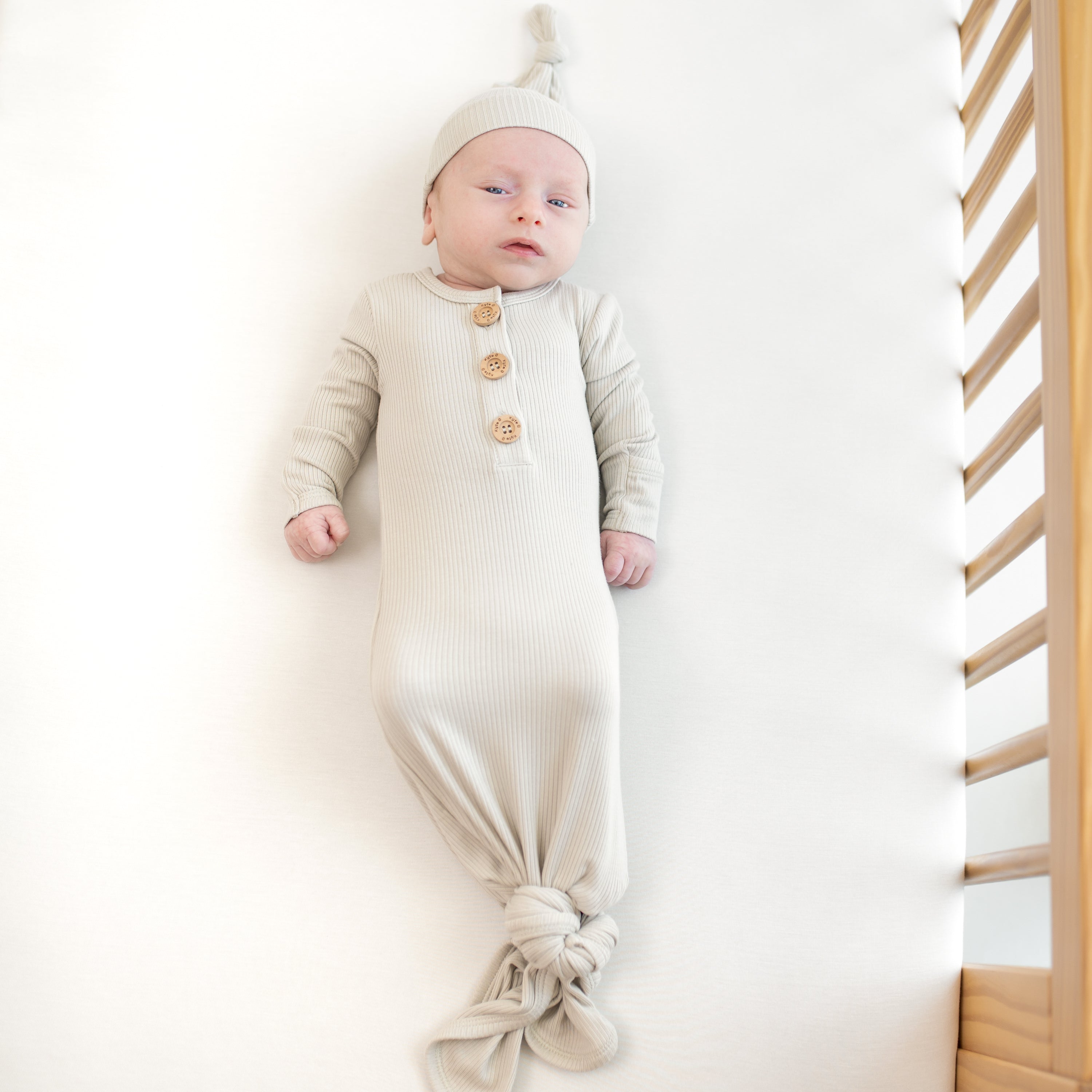 Baby in laying in a crib wearing the Kyte Baby Ribbed Knotted Gown with matching hat in the color oat.