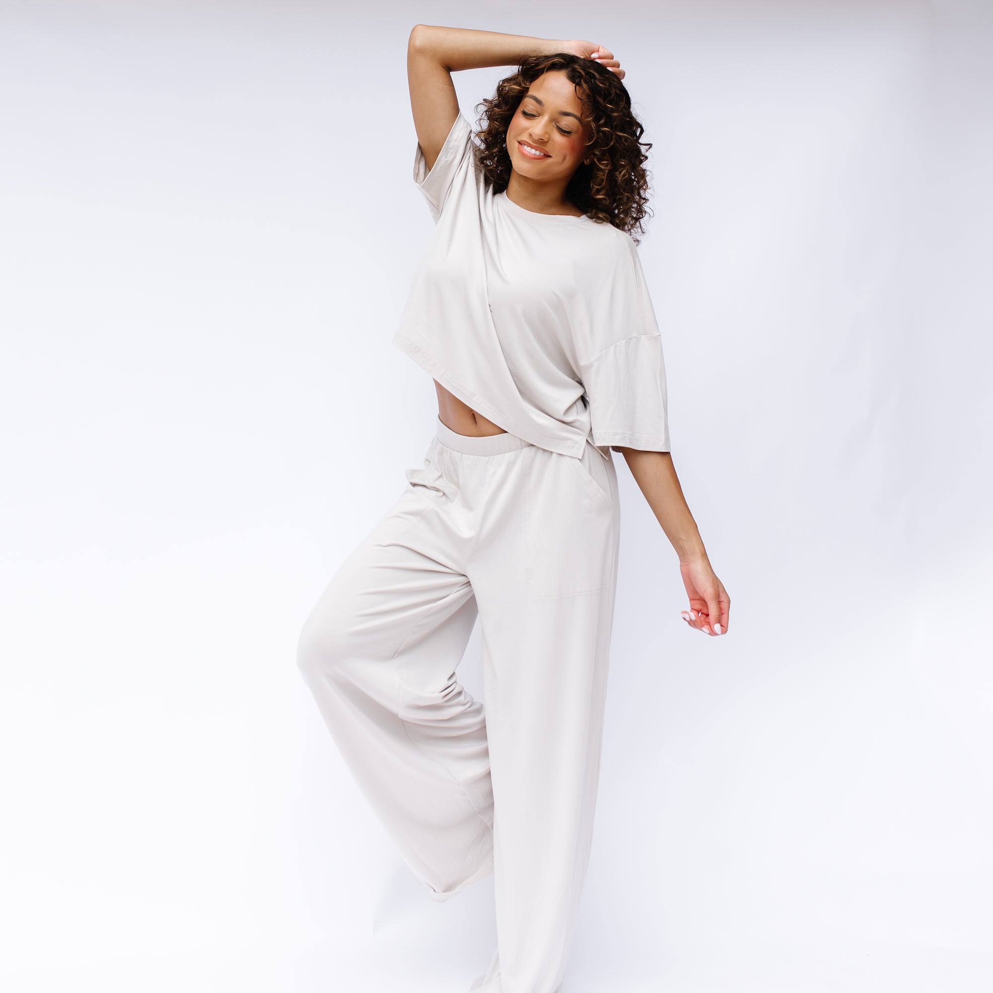 Female model standing with her eyes closed with one leg up and bent and one hand on top of her head wearing the Women's Short Sleeve Tee in Oat and matching wide leg pants