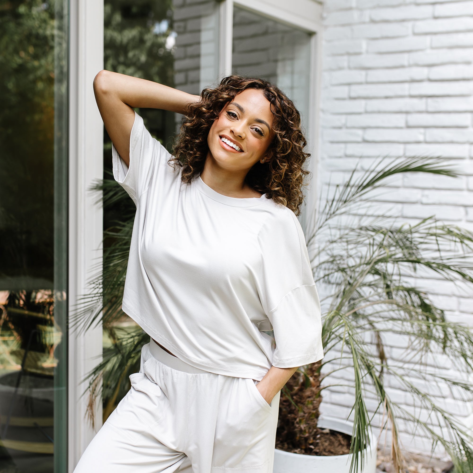 Smiling female model standing in front of a white brick house with one hand behind her head wearing the Women's Short Sleeve Tee in Oat and matching wide leg pants