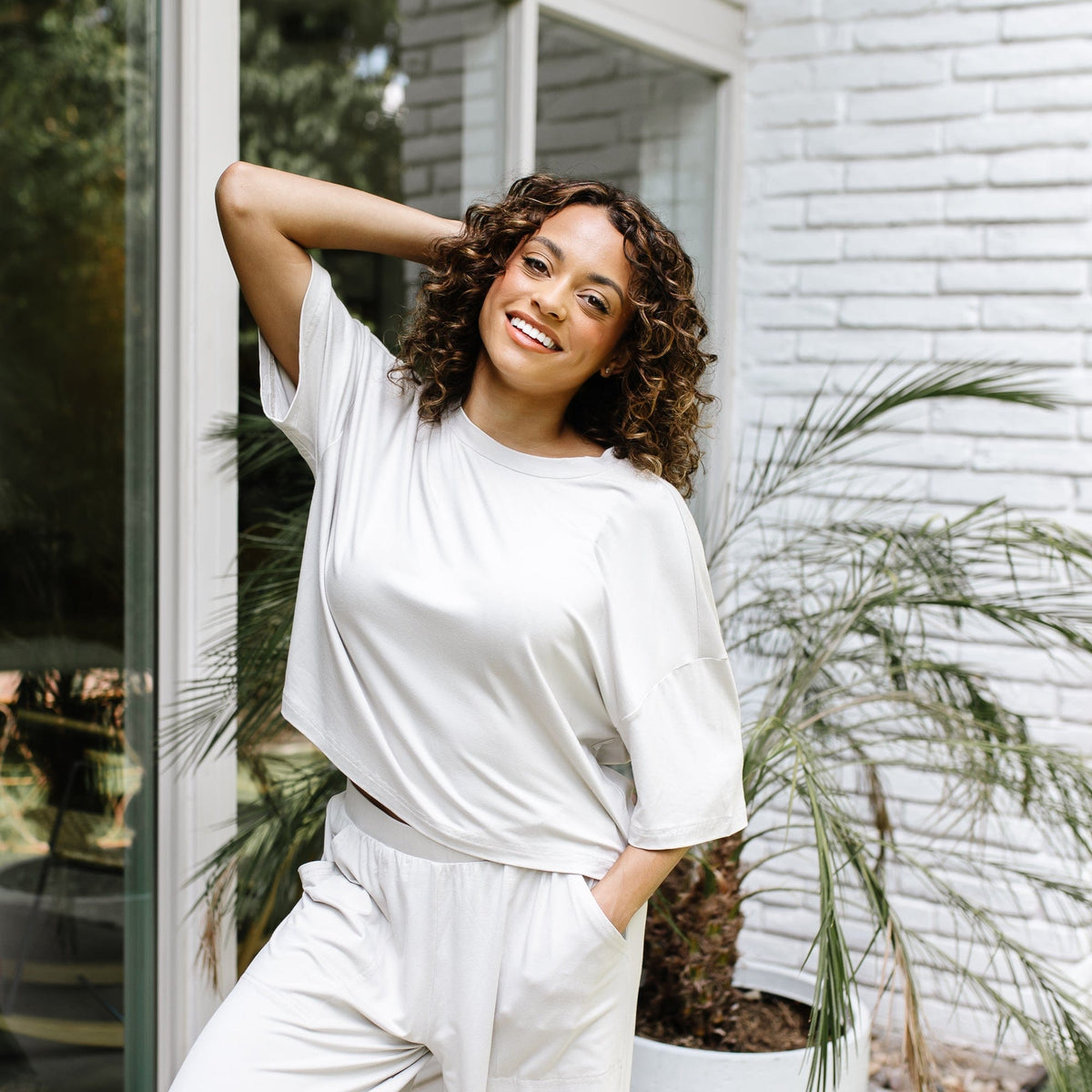 Smiling female model standing in front of a white brick house with one hand behind her head wearing the Women's Short Sleeve Tee in Oat and matching wide leg pants