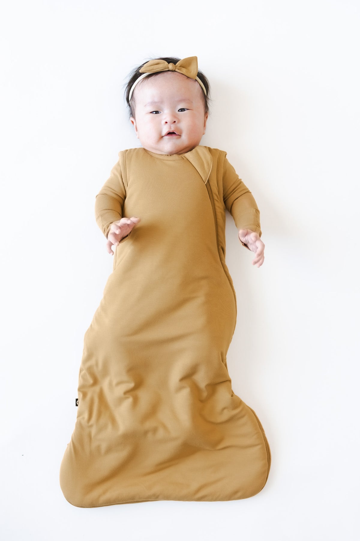 A baby lying on their back wearing a mustard yellow long-sleeve pajama and sleep sack and matching bow headband