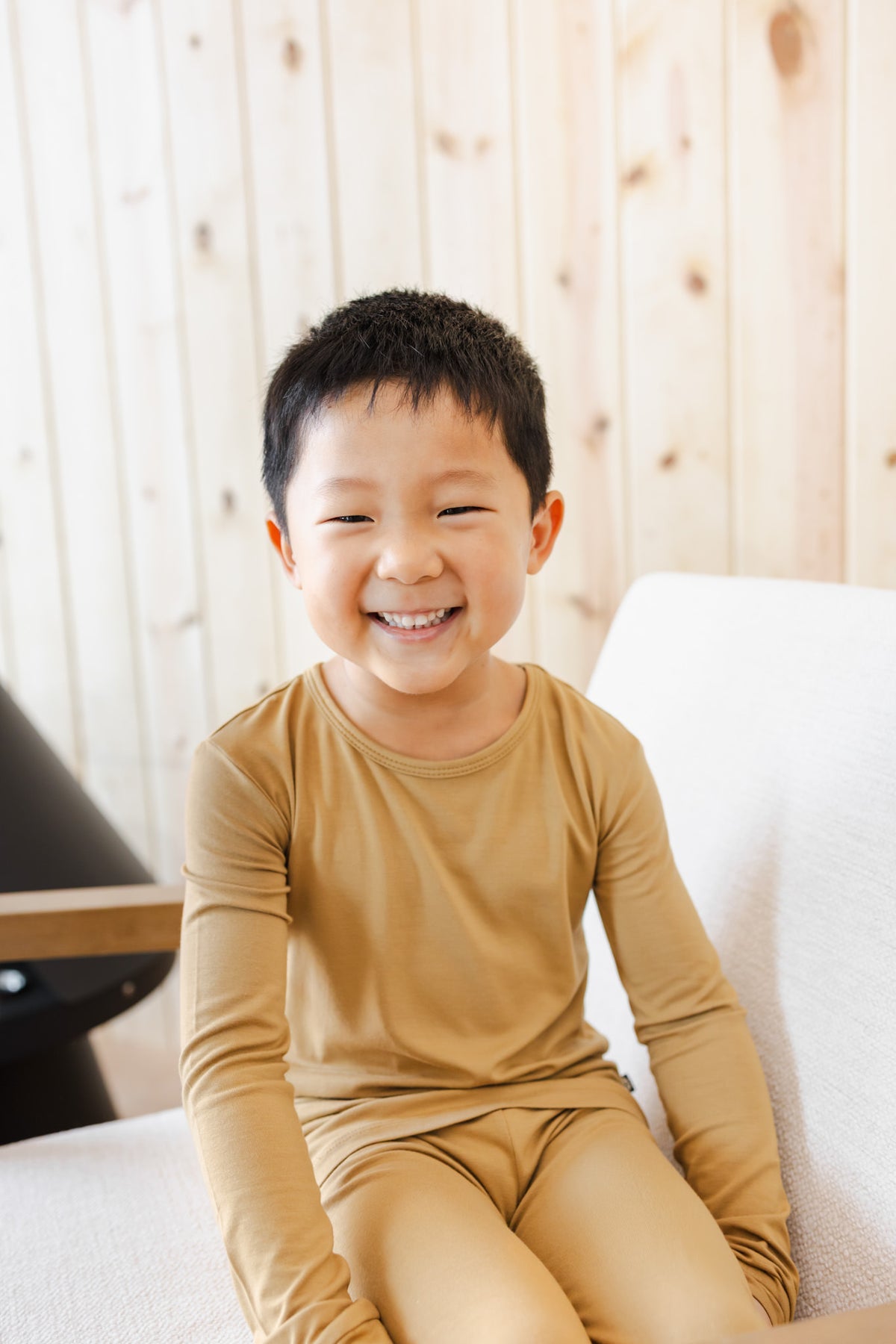 A young boy smiling while wearing a matching mustard yellow long-sleeve pajama set, sitting on a white sofa against a light wood-paneled wall.