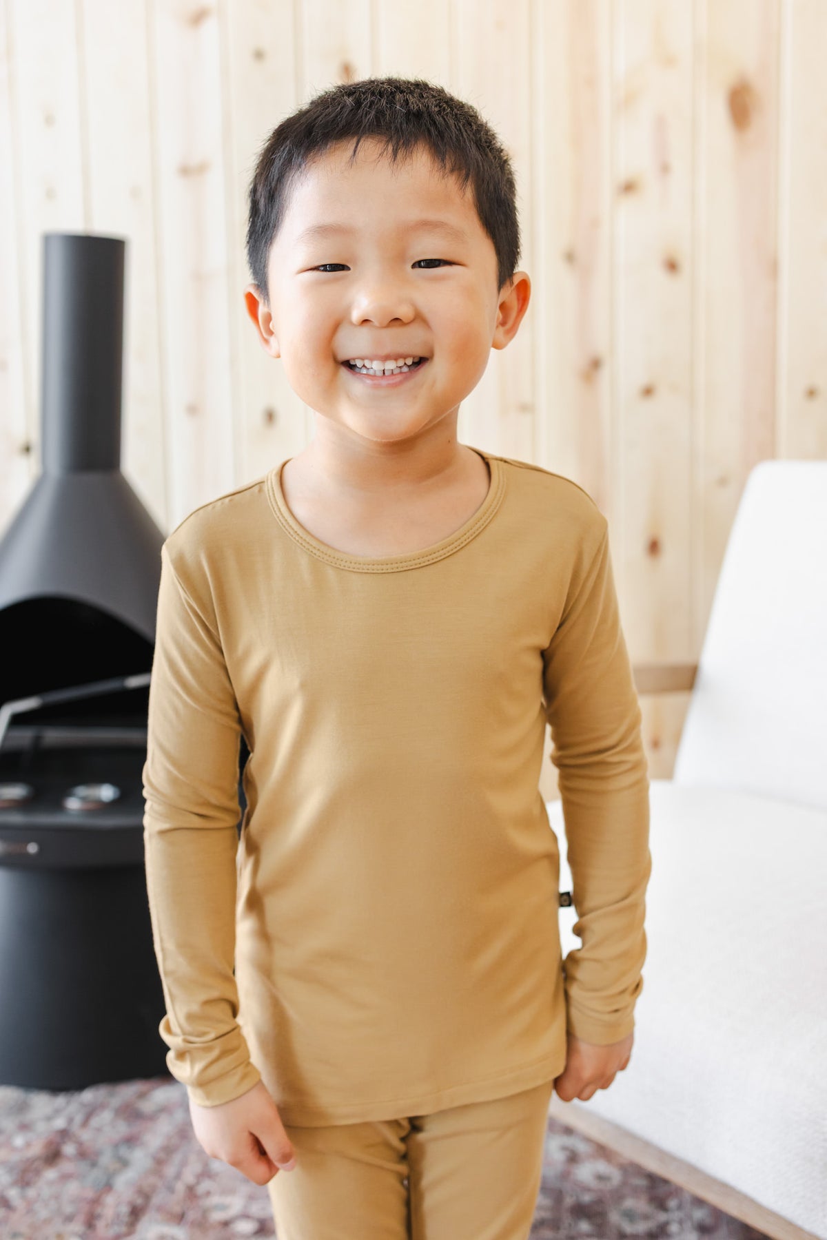 A young boy smiling and wearing a matching mustard pajama set, standing in a room with light wood-paneled walls.