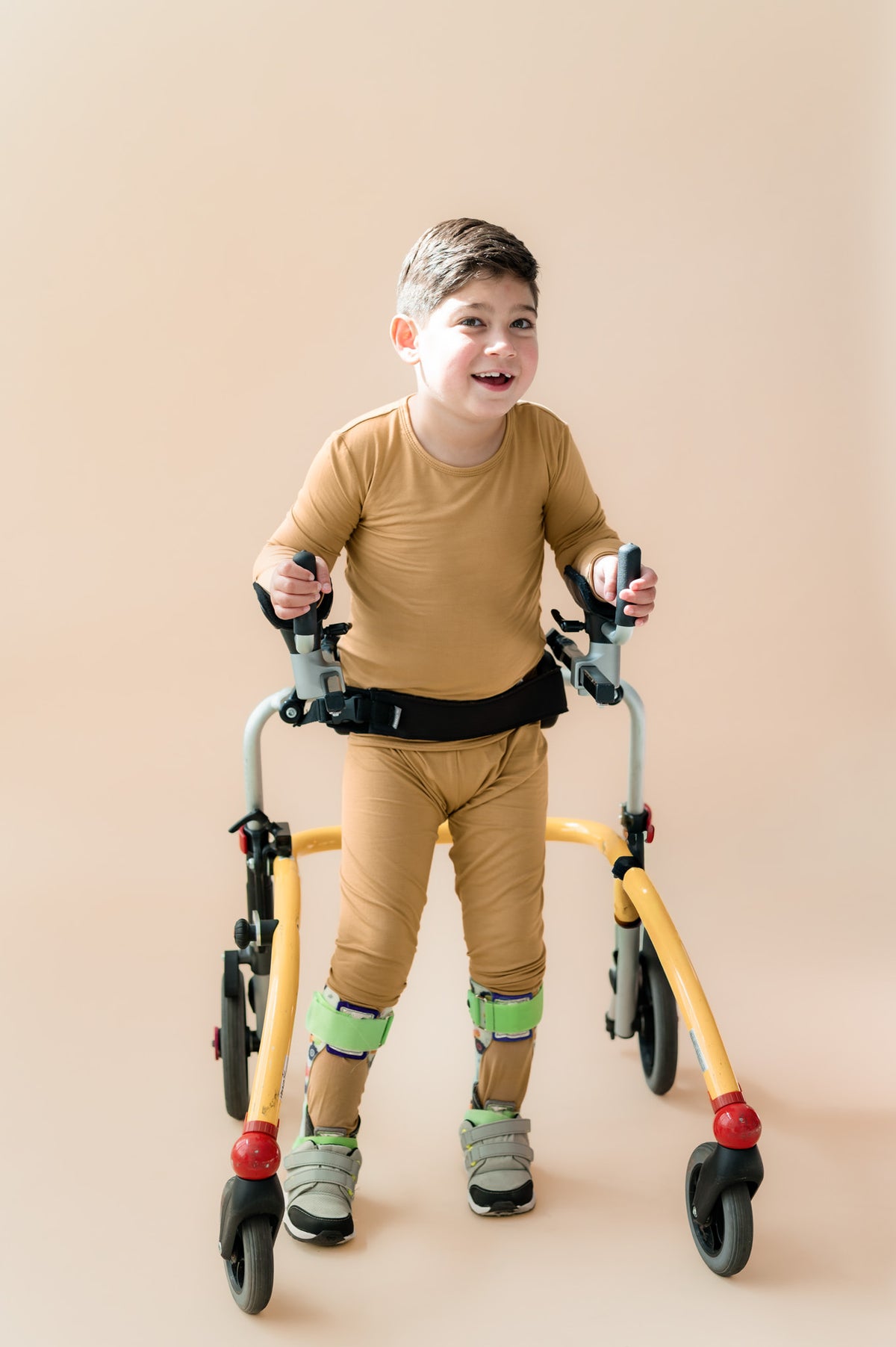 A young boy smiling and using a walker, wearing a mustard long-sleeve shirt and matching pajama set, standing against a solid beige background.
