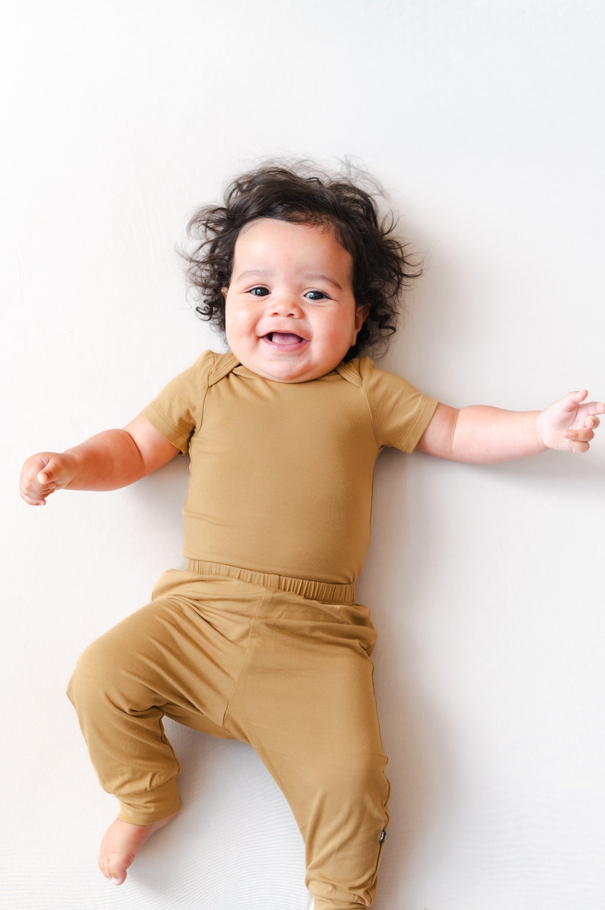 A smiling baby lying on their back wearing a matching short-sleeve bodysuit and jogger pants set in a warm mustard yellow color, set against a plain white background.