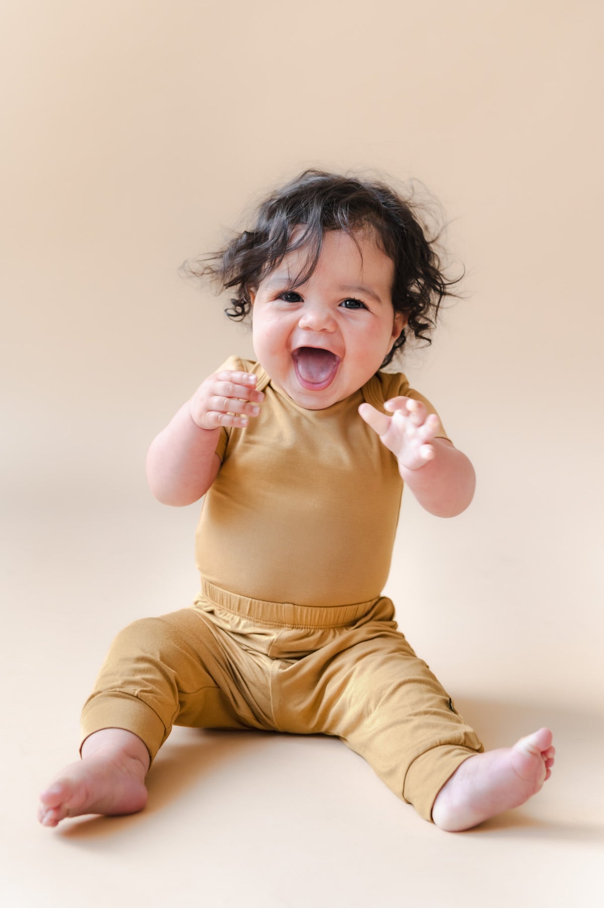 A smiling baby sitting on a warm beige studio background, wearing a matching mustard yellow bodysuit and pant