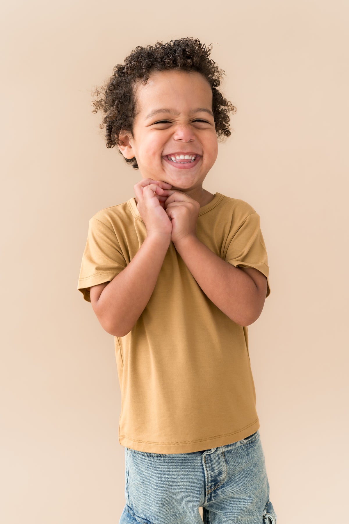 Boy wearing Toddler Basic Tee in Ochre over blue jeans on a beige background