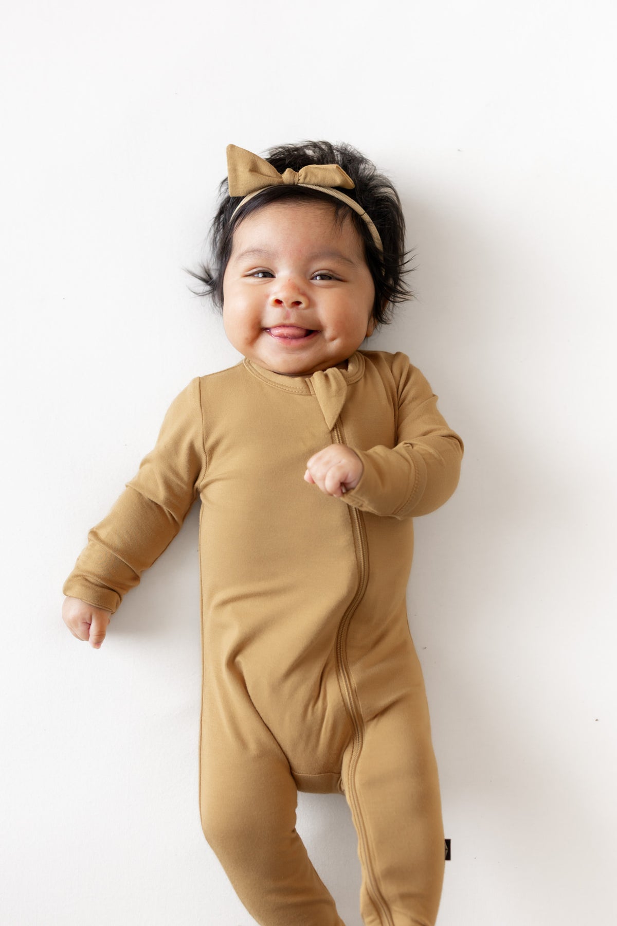 A smiling baby wearing a tan long-sleeve footed onesie and matching bow headband, lying on a white background.