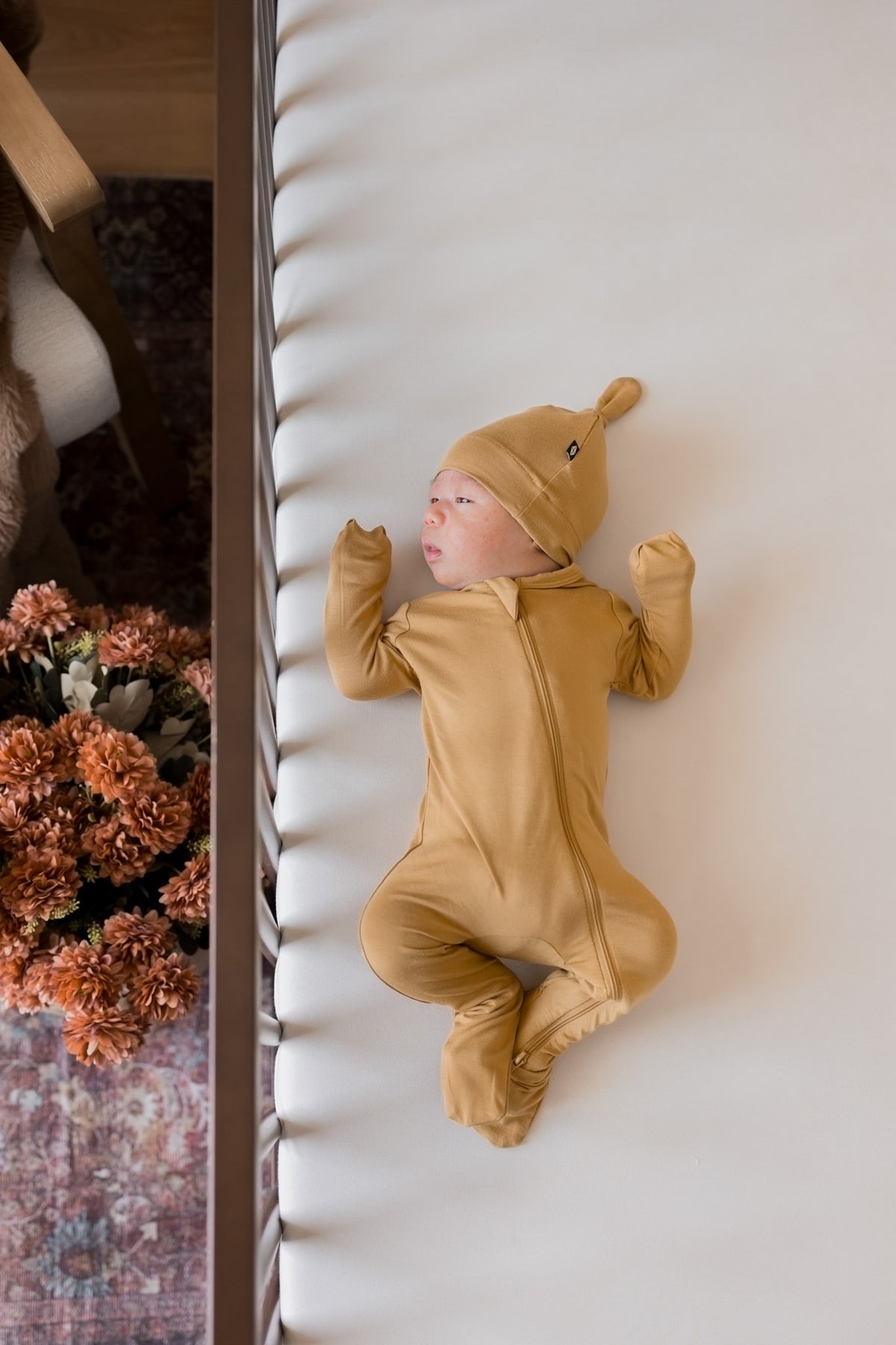A newborn baby wearing a mustard yellow full-body footed onesie and matching hat, lying on their back on a white textured blanket next to a wooden frame with dried flowers.