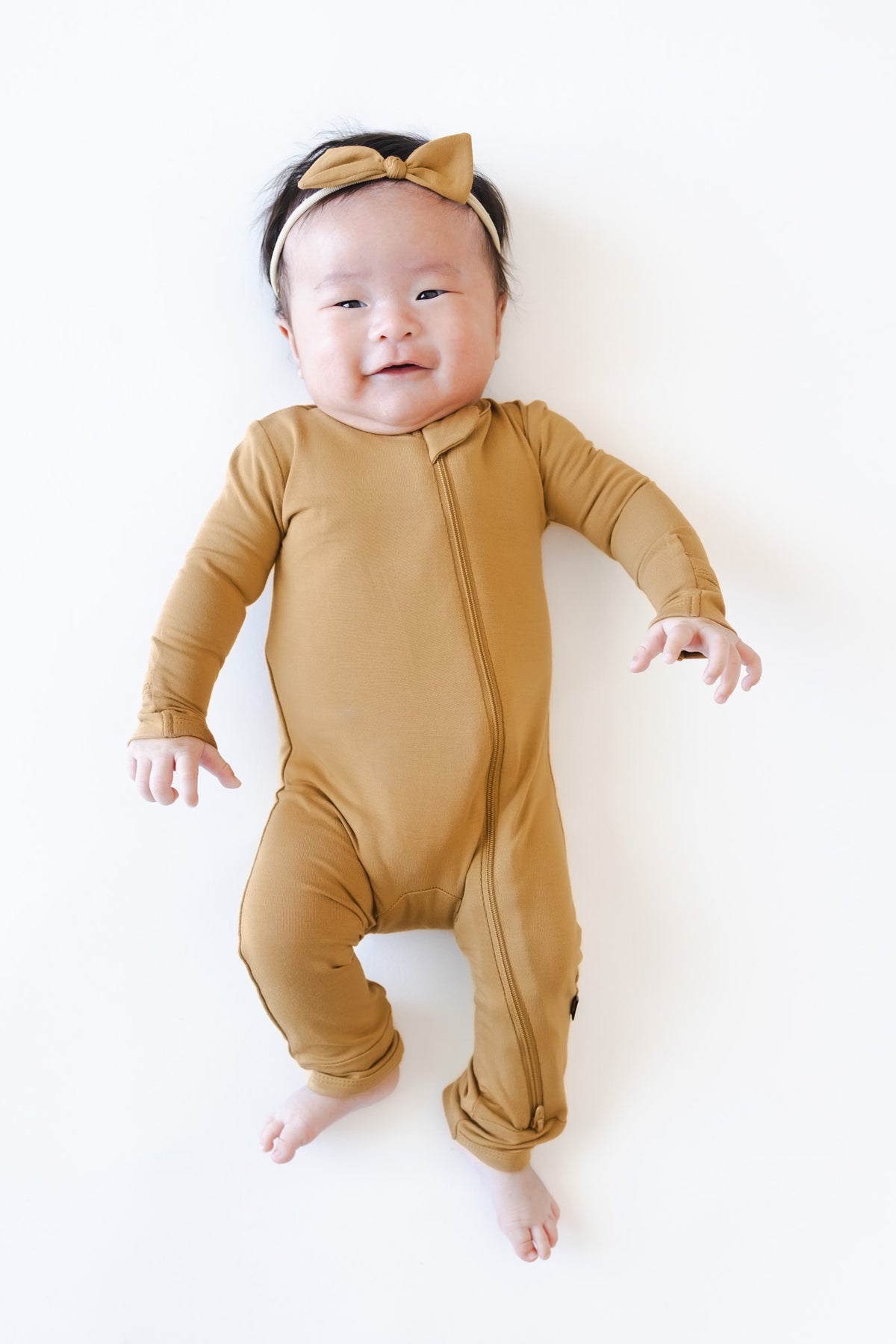 A baby lying on their back wearing a mustard yellow long-sleeve footless romper with a matching bow headband, photographed against a clean white background.
