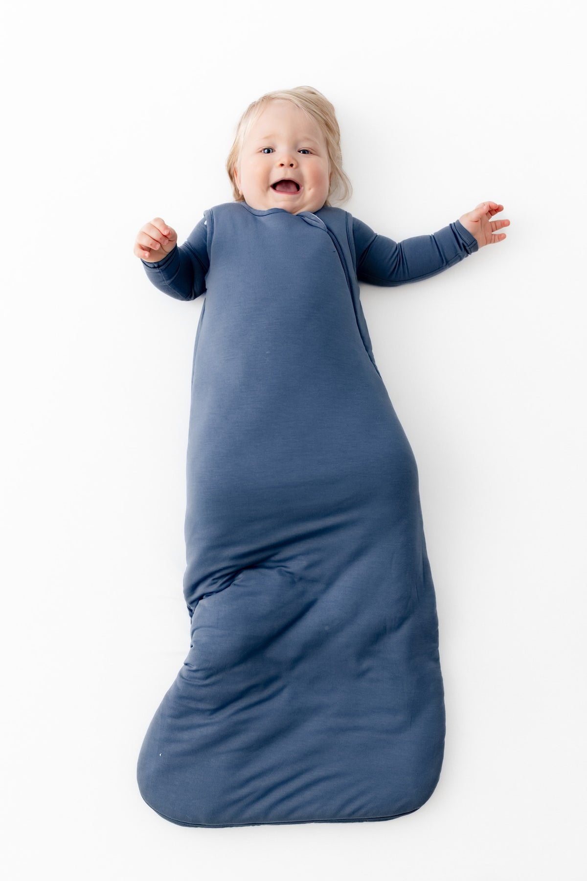 A baby lying on their back, smiling and laughing, wrapped in a blue sleeping bag against a solid white background.