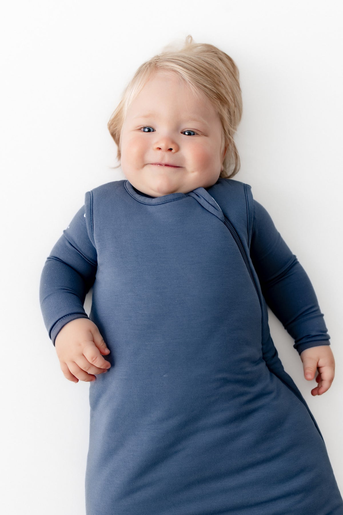 A baby lying on their back wearing a slate blue sleep sack against a clean white background.