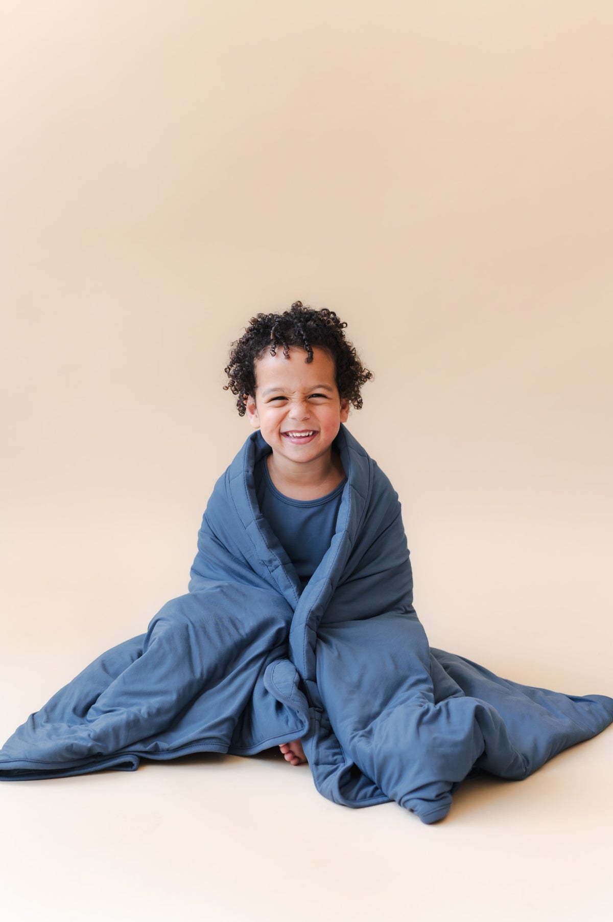 Boy smiling while wrapped in Youth Blanket in Oxford 2.5 on a beige background