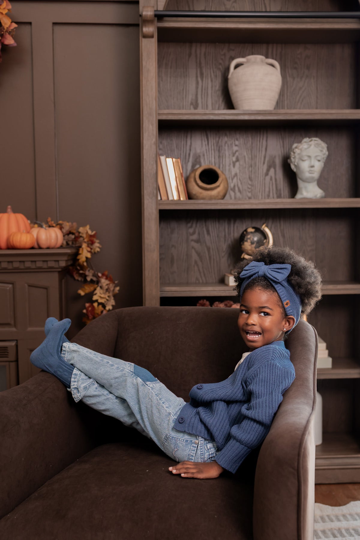 Girl lying on brown sofa wearing Chunky Knit Oversized Cardigan in Oxford over White T-shirt, Chunky knit bow headband, blue jeans and socks in oxford