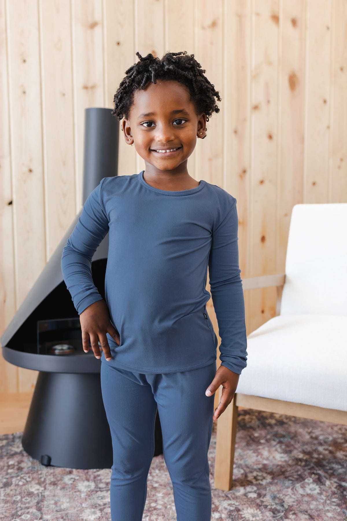 A young child wearing a matching slate blue long-sleeve pajama set, standing in a modern living room with a wood-paneled wall, a black fireplace, and a white chair.