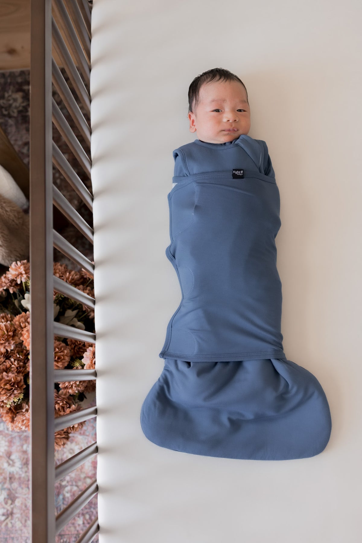 A newborn baby is lying on their back inside a crib, swaddled in a dusty slate blue sleep bag swaddler.