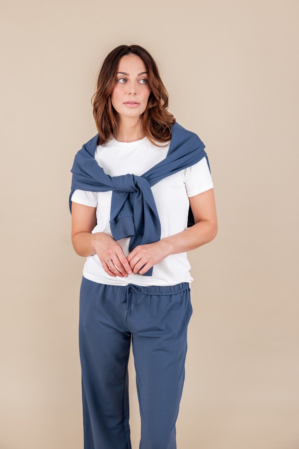 Woman wearing a white T-shirt and Women's Hoodie in Oxford color over her shoulders over similar color sweatpants on a beige background